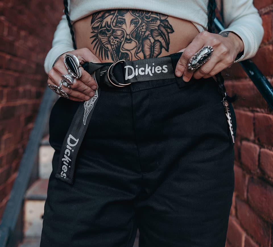 Dettaglio di persona con tatuaggio di leone sull'addome, che indossa pantaloni neri e una cintura Dickies con logo Harley-Davidson. Le mani con anelli tengono la cintura.