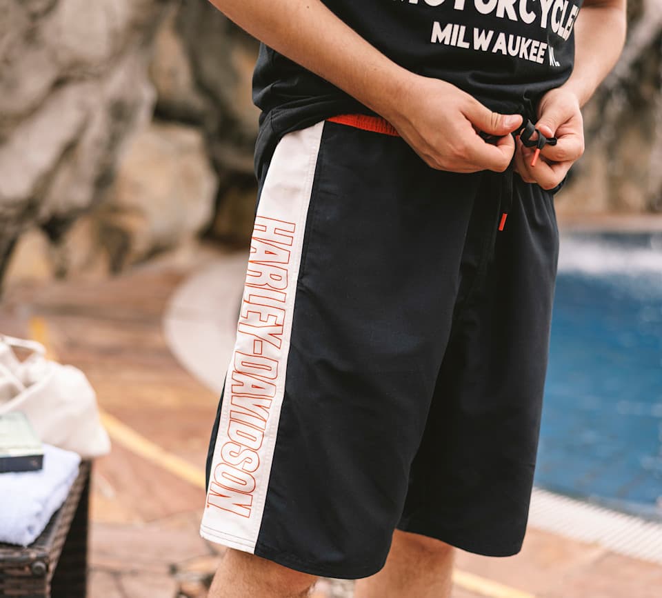 Persona che indossa pantaloncini da bagno neri con banda laterale bianca e logo Harley-Davidson arancione, mentre stringe il cordino in vita a bordo piscina.