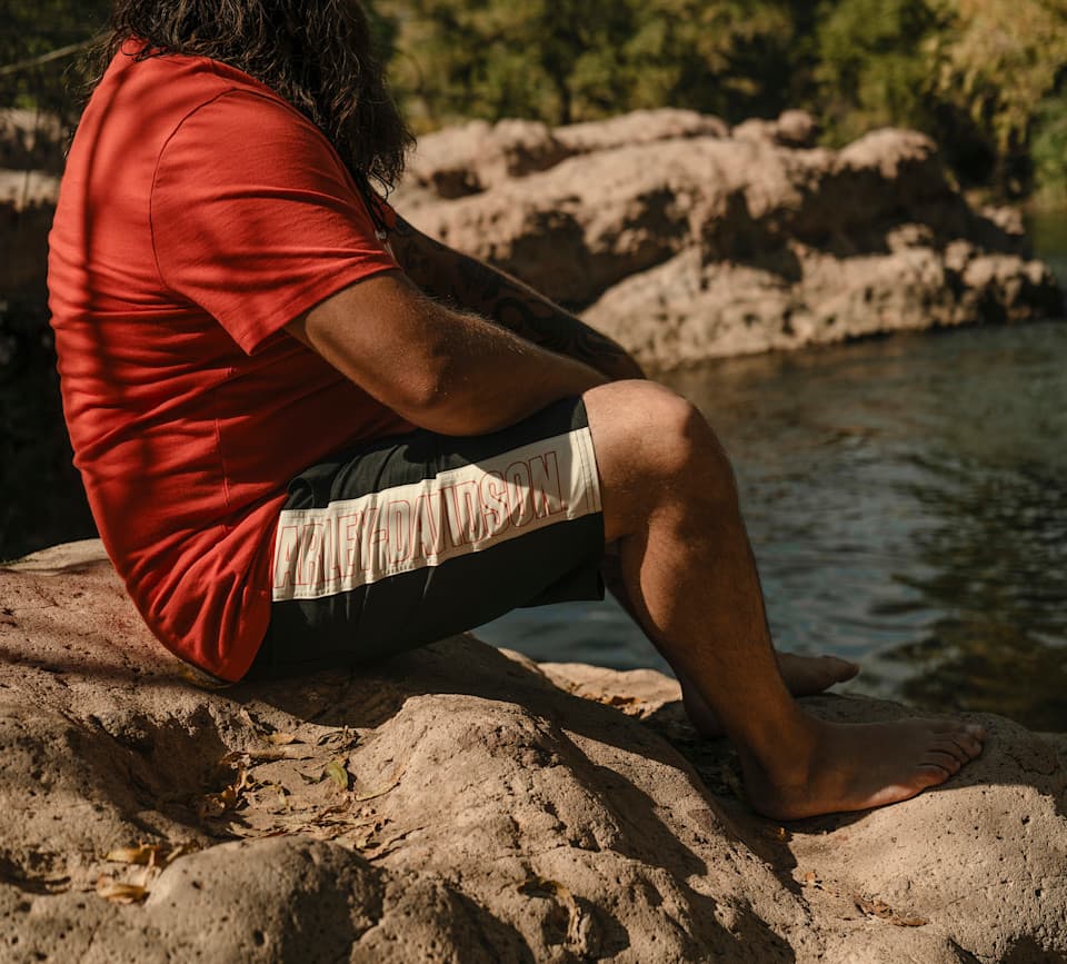 Uomo con abbigliamento Harley-Davidson, shorts neri con banda chiara e logo rosso, seduto su rocce in ambiente naturale.