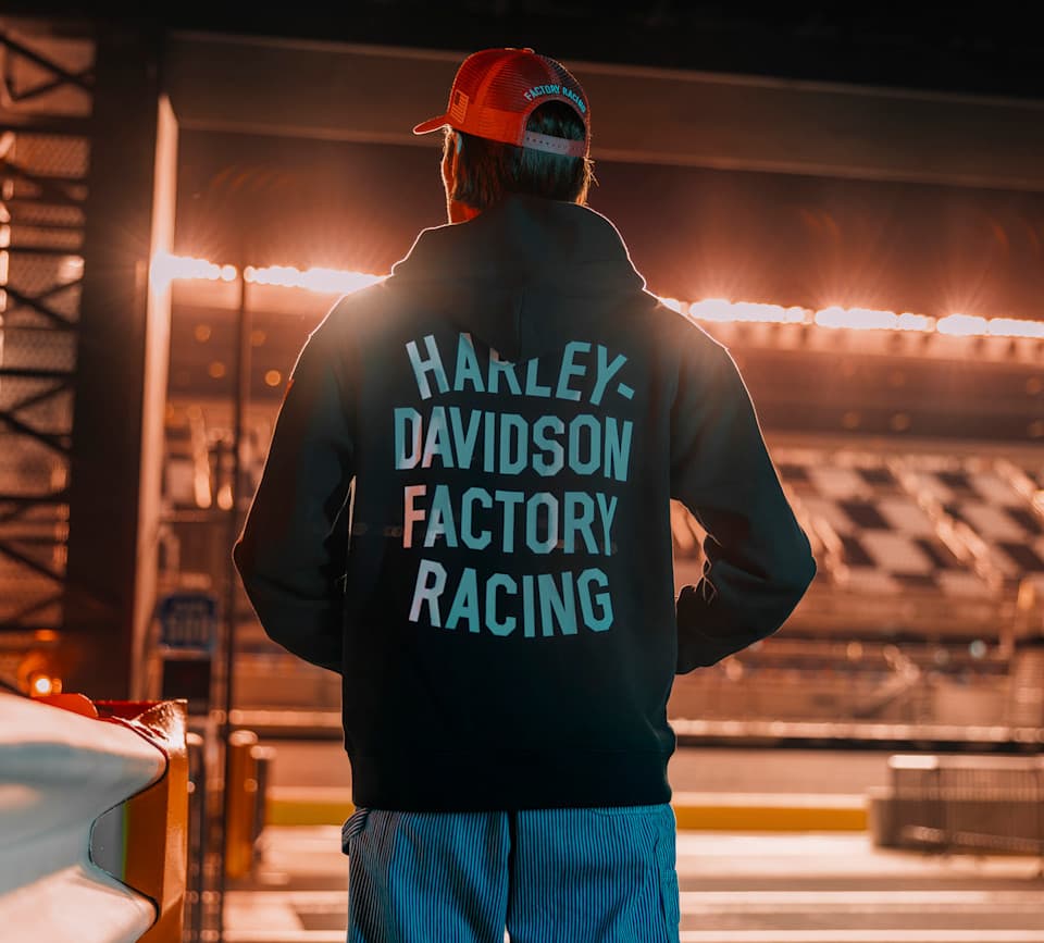 Vista posteriore di persona con felpa nera e cappellino rosso, entrambi con stampa 'HARLEY-DAVIDSON FACTORY RACING', in ambiente sportivo illuminato.