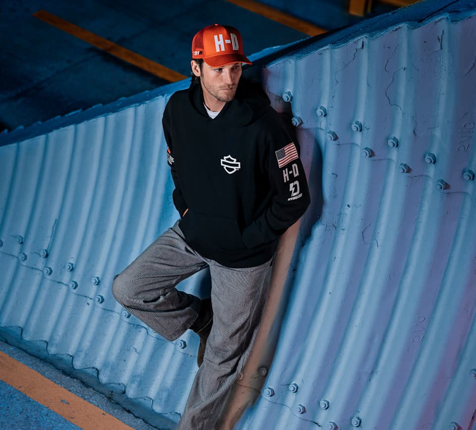 Uomo indossa cappellino trucker arancione con logo H-D e felpa nera con scudo Harley-Davidson e patch bandiera USA sulla manica, appoggiato a una parete strutturale blu.
