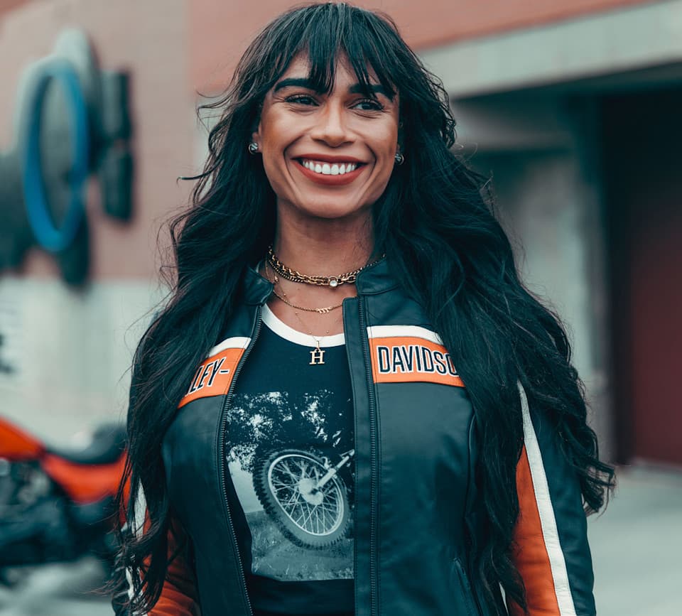 Donna sorridente con giacca in pelle Harley-Davidson nera, strisce arancioni e bianche, e maglietta con grafica moto. Sfondo sfocato con moto.