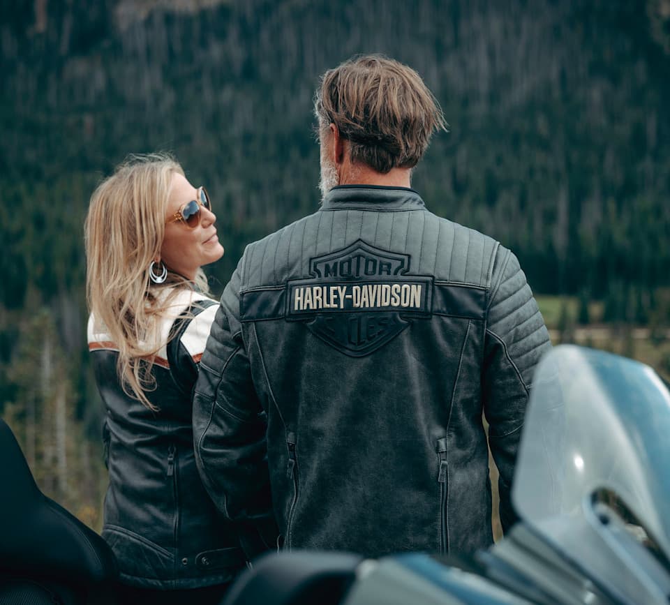 Coppia con giacche in pelle Harley-Davidson, schiena rivolta, ammira paesaggio montano. Logo Harley-Davidson sulla giacca dell'uomo. Moto in primo piano.