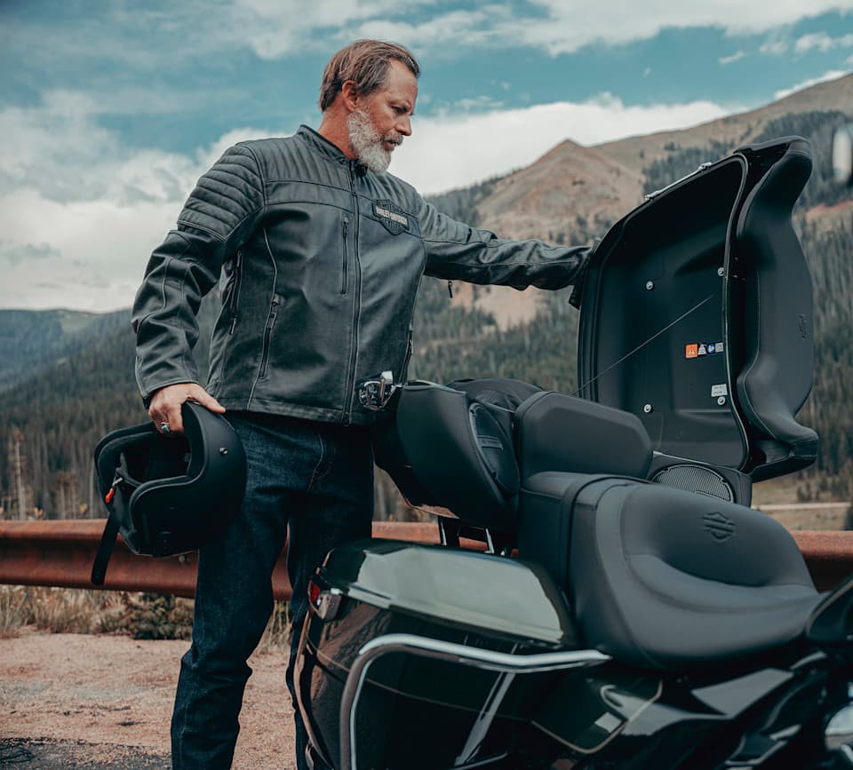 Motociclista con giacca in pelle Harley-Davidson e casco, apre il top case di una moto touring in un paesaggio montano.