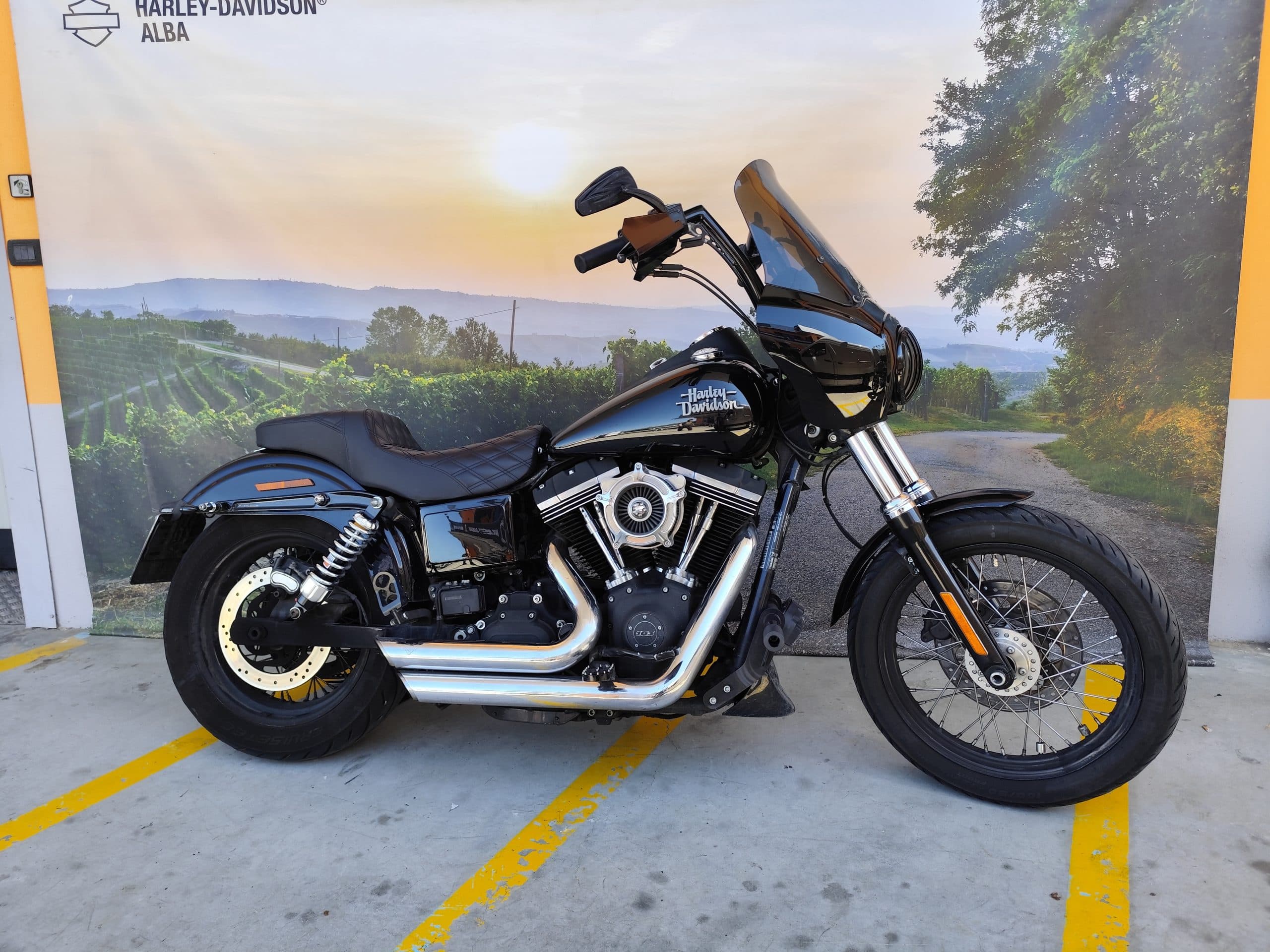 Motocicletta Harley-Davidson Dyna nera, motore 103, scarichi cromati, esposta presso la concessionaria Harley-Davidson Alba con sfondo paesaggistico.