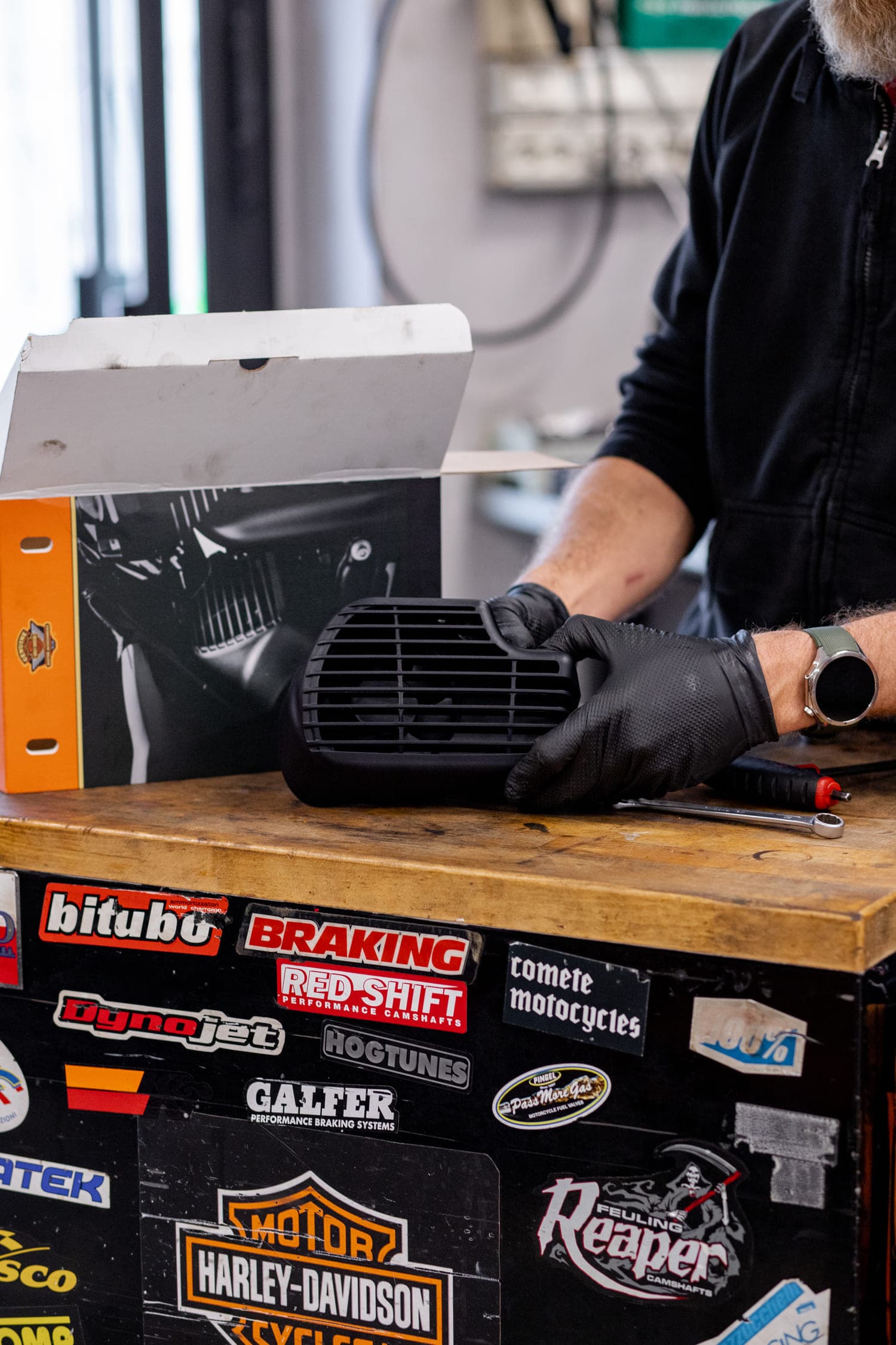 Installazione accessori e ricambi Harley-Davidson Componente motore Harley-Davidson in mano a meccanico guantato su banco da lavoro. Scatola originale e attrezzi in officina specializzata.