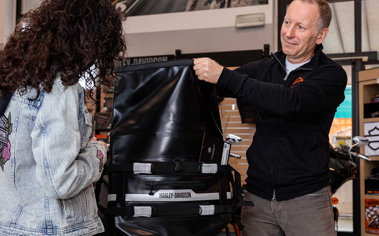 Staff mostra borsa moto Harley-Davidson a cliente Addetto Harley-Davidson mostra una borsa da moto impermeabile nera con logo a una cliente, illustrando le funzionalità in concessionaria.