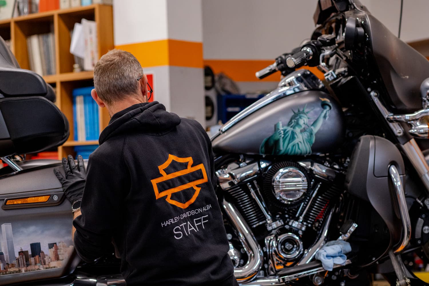 Harley-Davidson Alba: personalizzazione moto con dettagli unici Personale Harley-Davidson Alba in felpa staff esegue manutenzione su moto customizzata con aerografie Statua della Libertà e skyline.