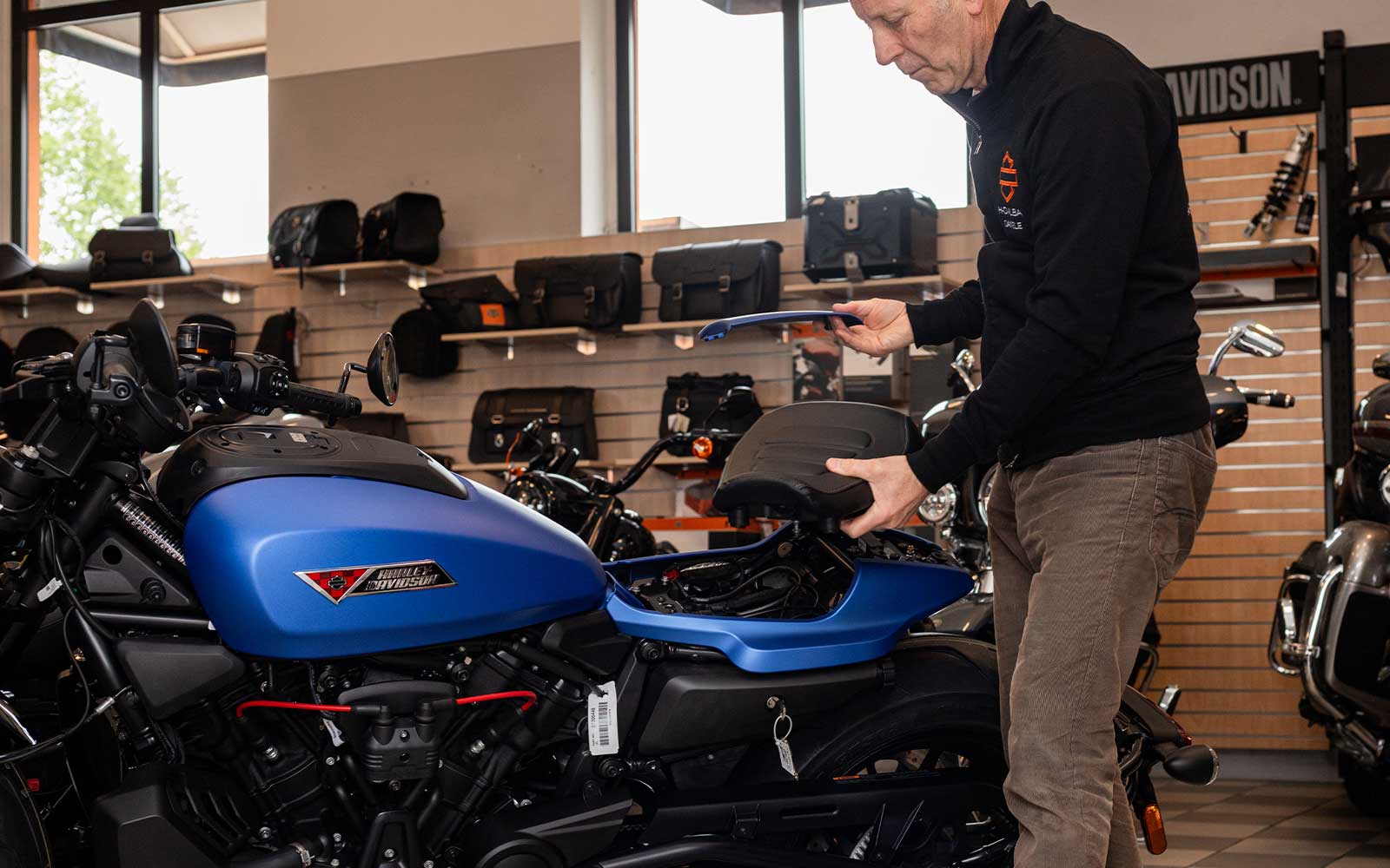 Tecnico Harley-Davidson personalizza moto Sportster S Tecnico Harley-Davidson in concessionaria mentre maneggia la sella e un componente blu di una moto, con accessori esposti sullo sfondo.