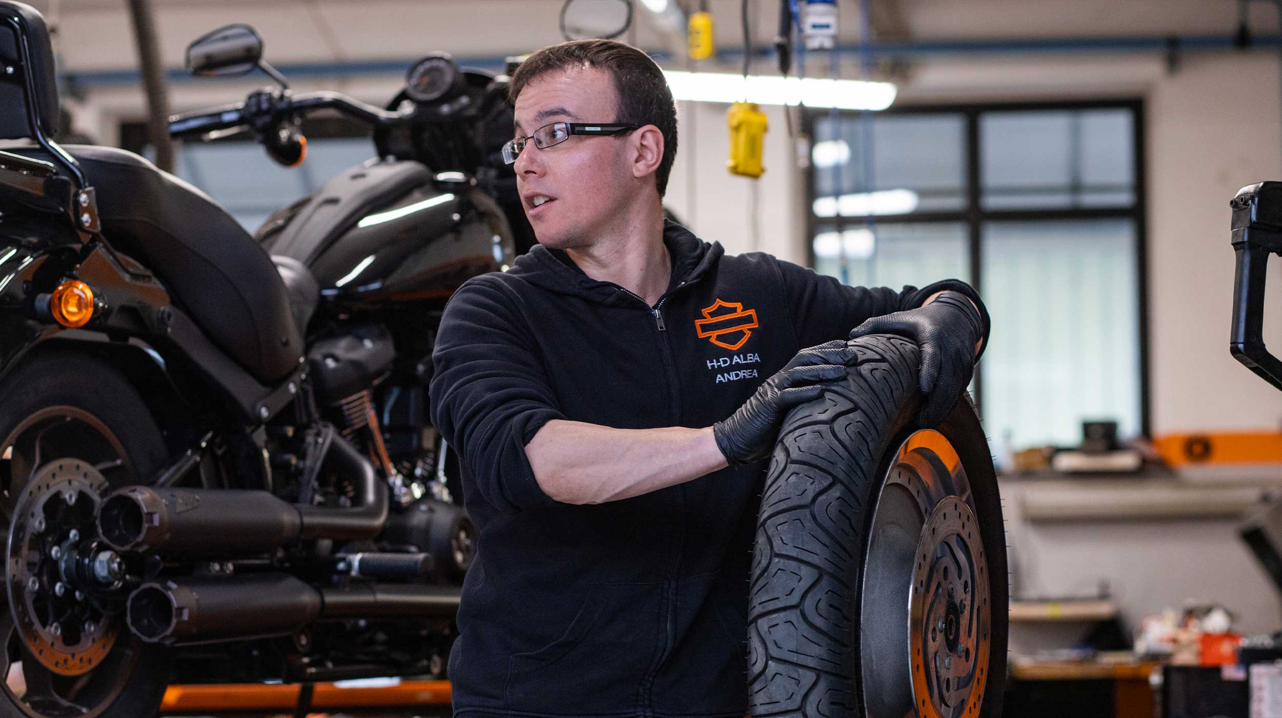 Andrea di H-D Alba in officina con ruota moto Harley-Davidson Meccanico Harley-Davidson Alba, Andrea, con guanti neri, tiene una ruota di moto con pneumatico, in officina.