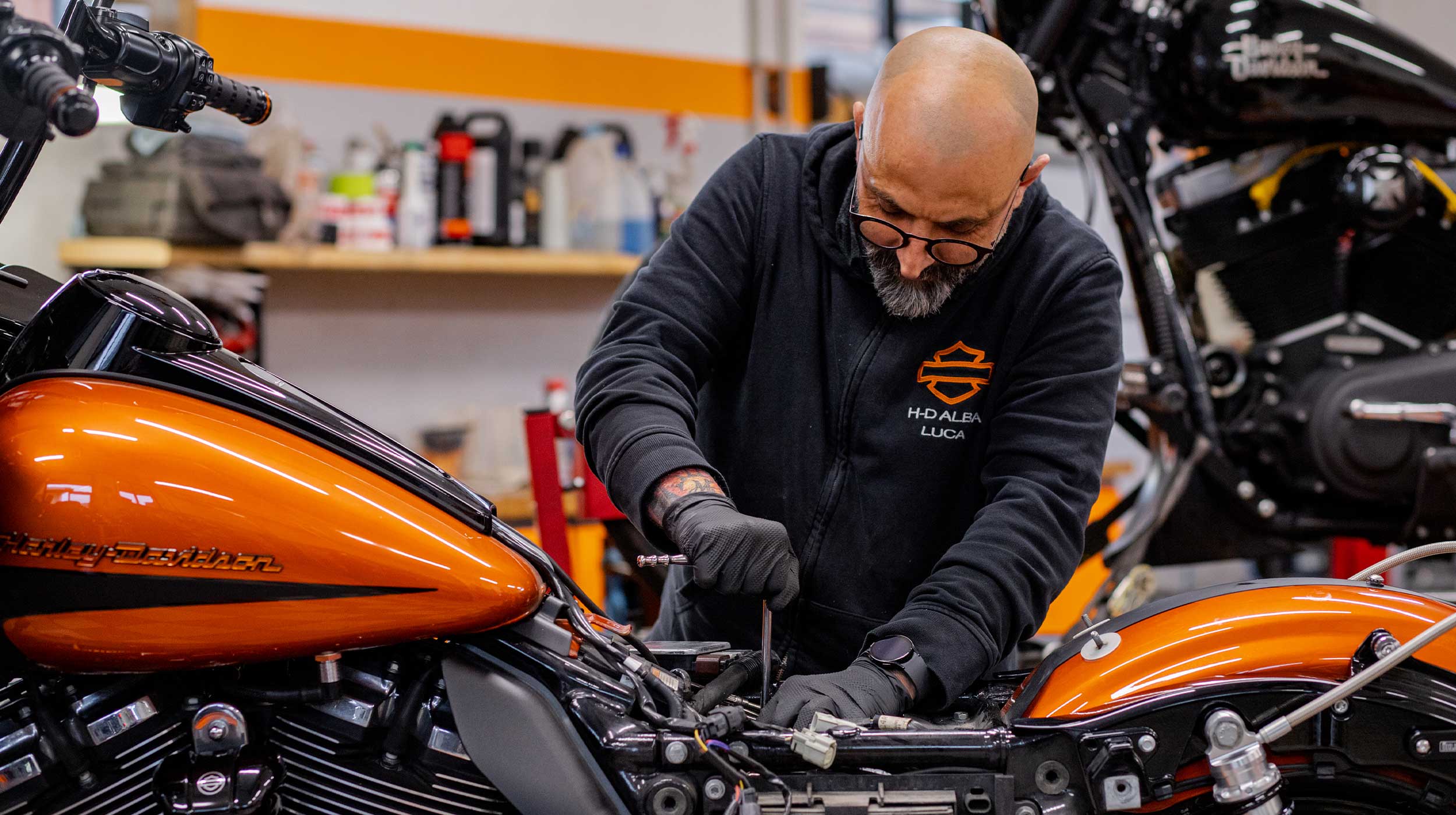Officina Harley-Davidson Alba: Luca al lavoro Meccanico Harley-Davidson di H-D Alba Luca esegue manutenzione su una moto arancione in officina, concentrato sui motori.