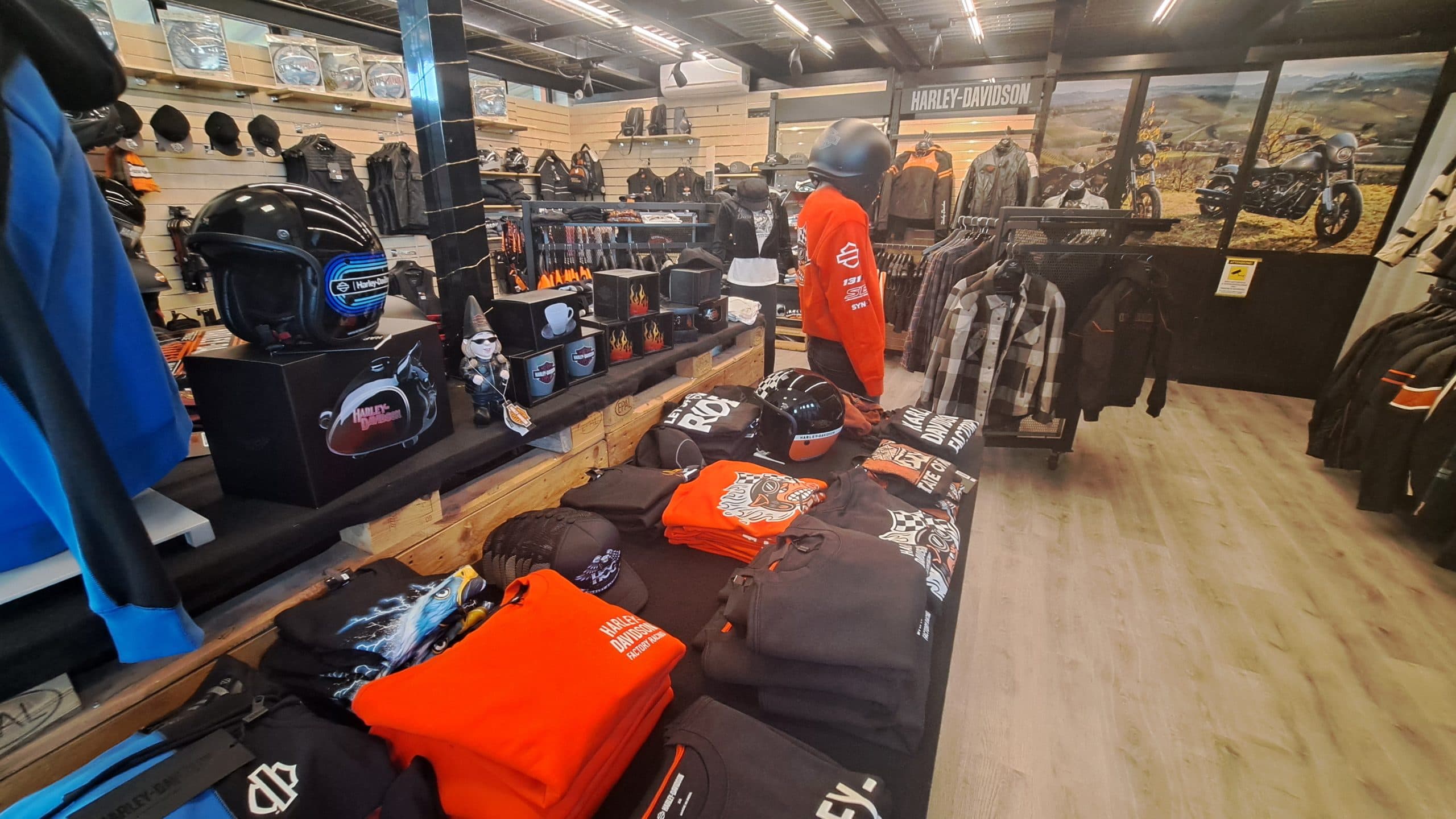 1000813333-scaled Ambiente retail Harley-Davidson con esposizione di abbigliamento, caschi e accessori. In primo piano, magliette, felpe e caschi brandizzati.