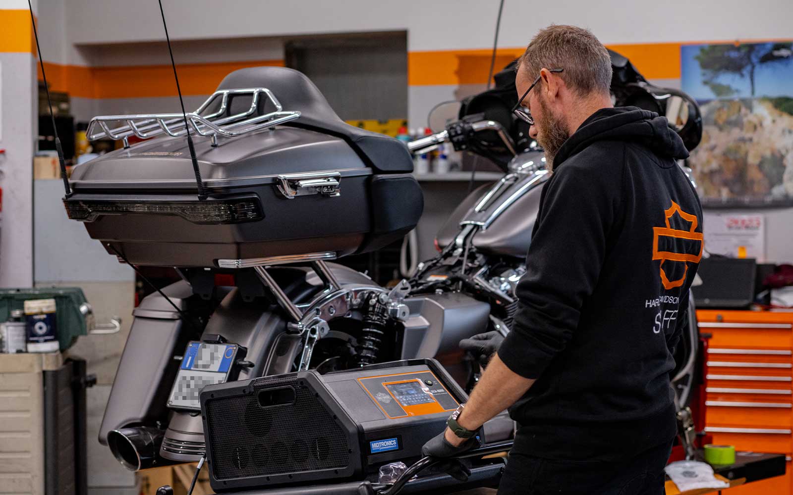 Manutenzione e diagnostica Harley-Davidson in officina Meccanico Harley-Davidson con felpa logata opera un dispositivo diagnostico Midtronics su una moto touring in officina.
