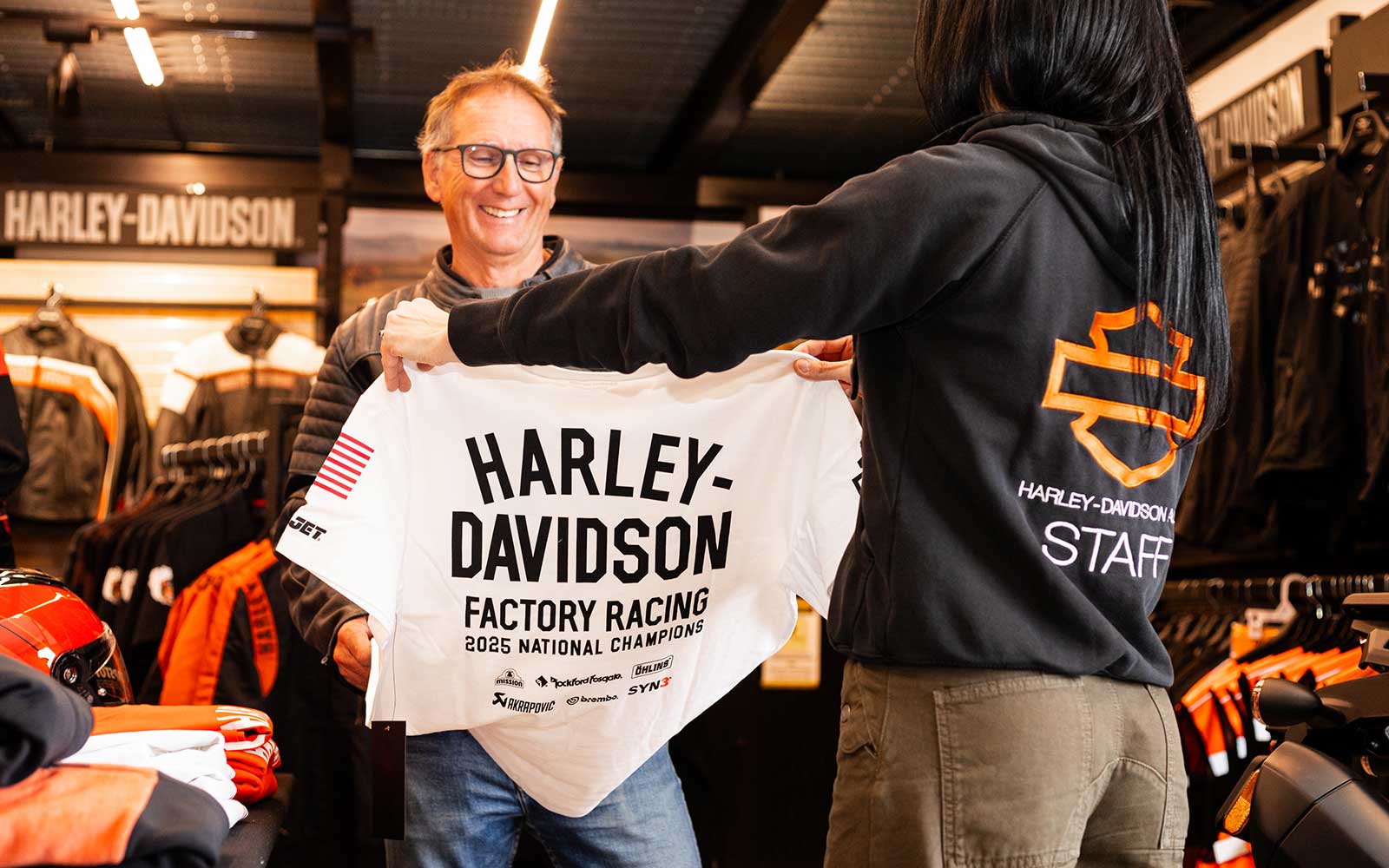 Cliente felice con maglietta Harley-Davidson Racing 2025 Cliente sorridente osserva una maglietta Harley-Davidson Factory Racing 2025 National Champions, mostrata da un membro dello staff in un negozio Harley-Davidson.