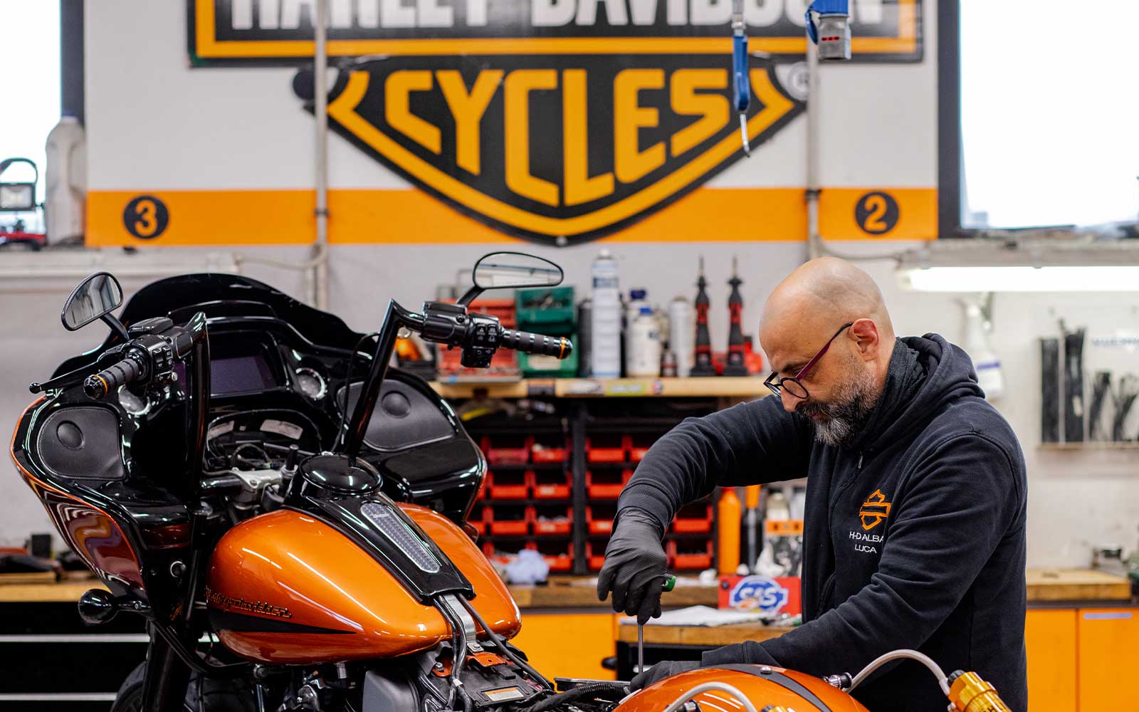 Meccanico Harley-Davidson al lavoro nell’officina di Alba Meccanico Harley-Davidson di Alba in officina, con felpa logata, esegue manutenzione su moto touring arancione.