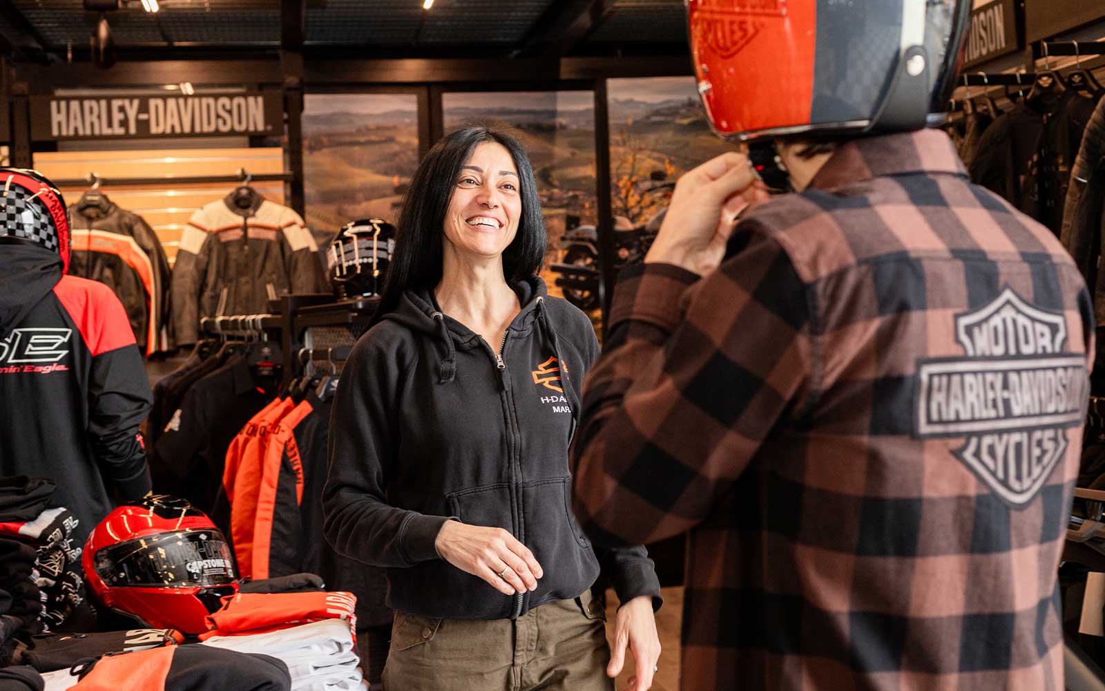 Vivi l’esperienza Harley-Davidson: abbigliamento e caschi Cliente prova casco e abbigliamento Harley-Davidson in concessionaria. La commessa sorride, sullo sfondo giacche e caschi per moto.