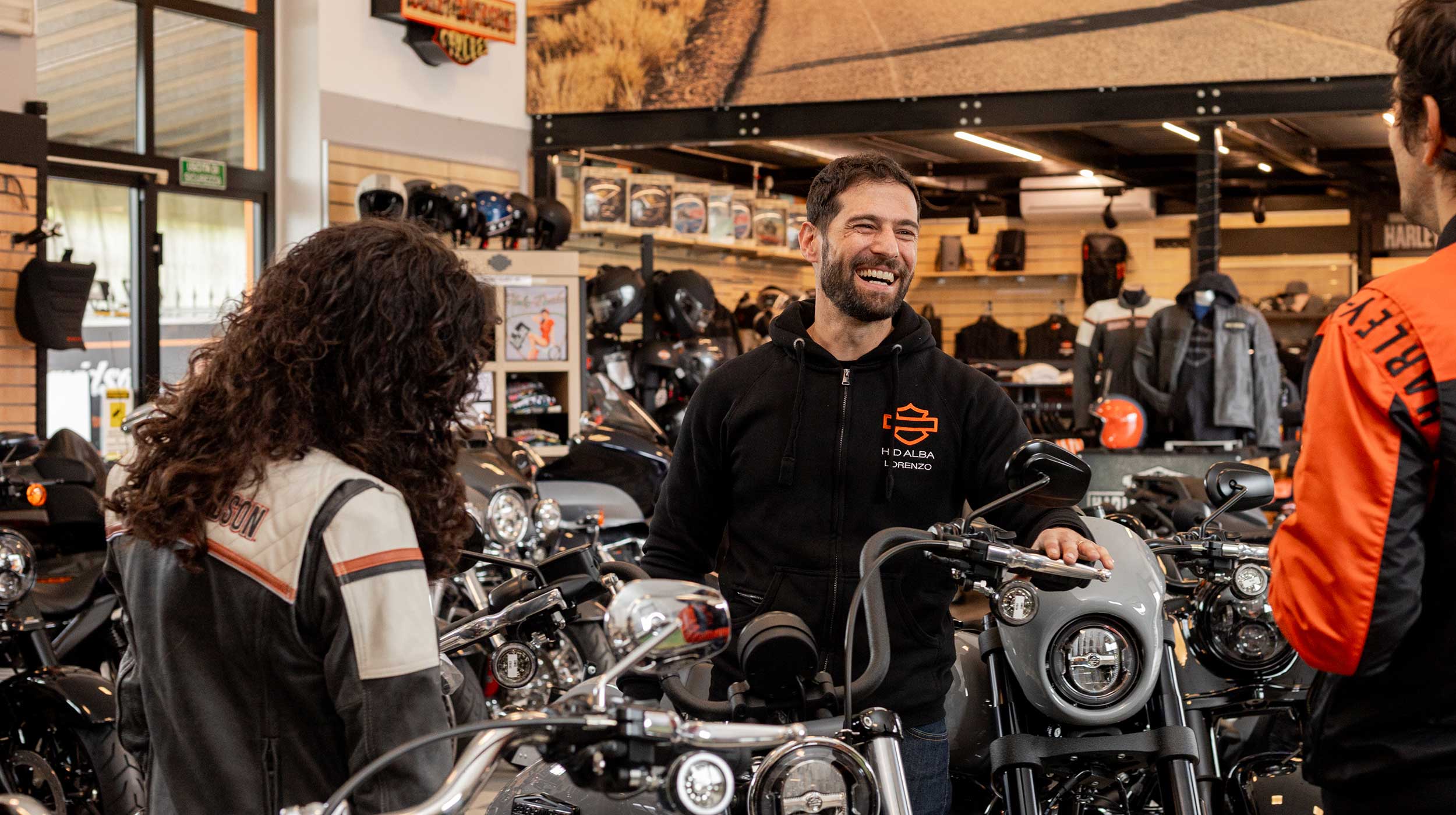 Clienti e venditore Harley-Davidson ad Alba Addetto Harley-Davidson Alba sorride, assistendo clienti tra modelli di moto e abbigliamento tecnico in esposizione.