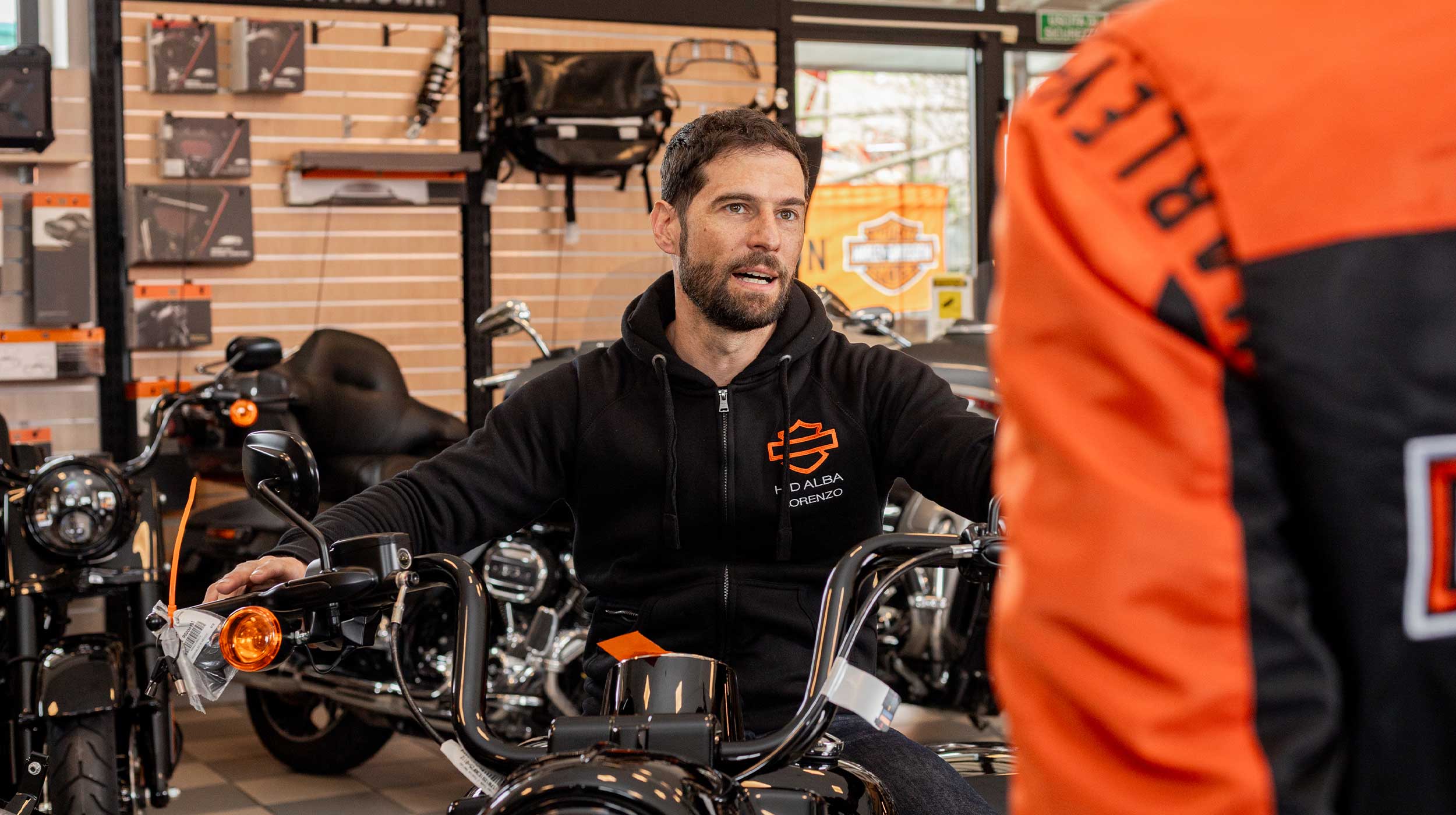 Consulenza e prova moto Harley-Davidson in concessionaria Alba Uomo con felpa nera Harley-Davidson con logo arancione e scritta "D'ALBA LORENZO" seduto su una moto nera, mentre parla con una persona di spalle con giacca arancione e nera. Sullo sfondo, scaffali con accessori e ricambi Harley-Davidson e un banner con il logo del marchio.
