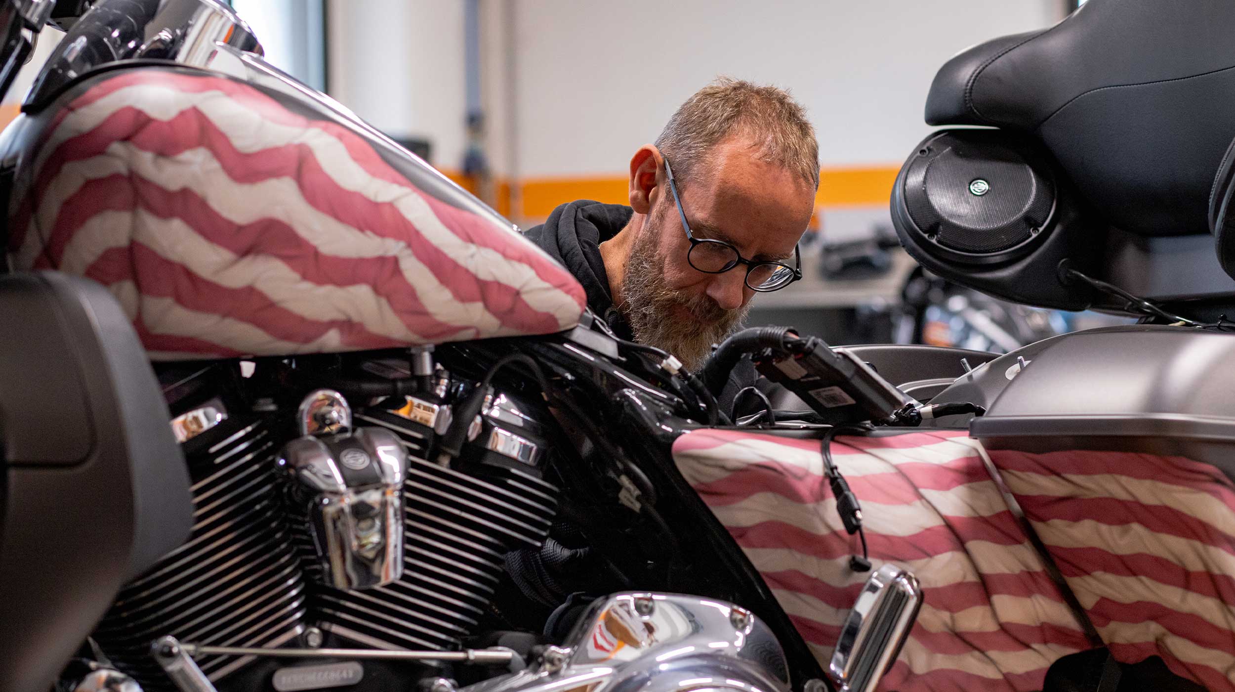 Meccanico Harley-Davidson al lavoro su moto custom Meccanico con barba e occhiali che lavora su una moto Harley-Davidson con serbatoio decorato con bandiera americana, concentrato sui cablaggi.