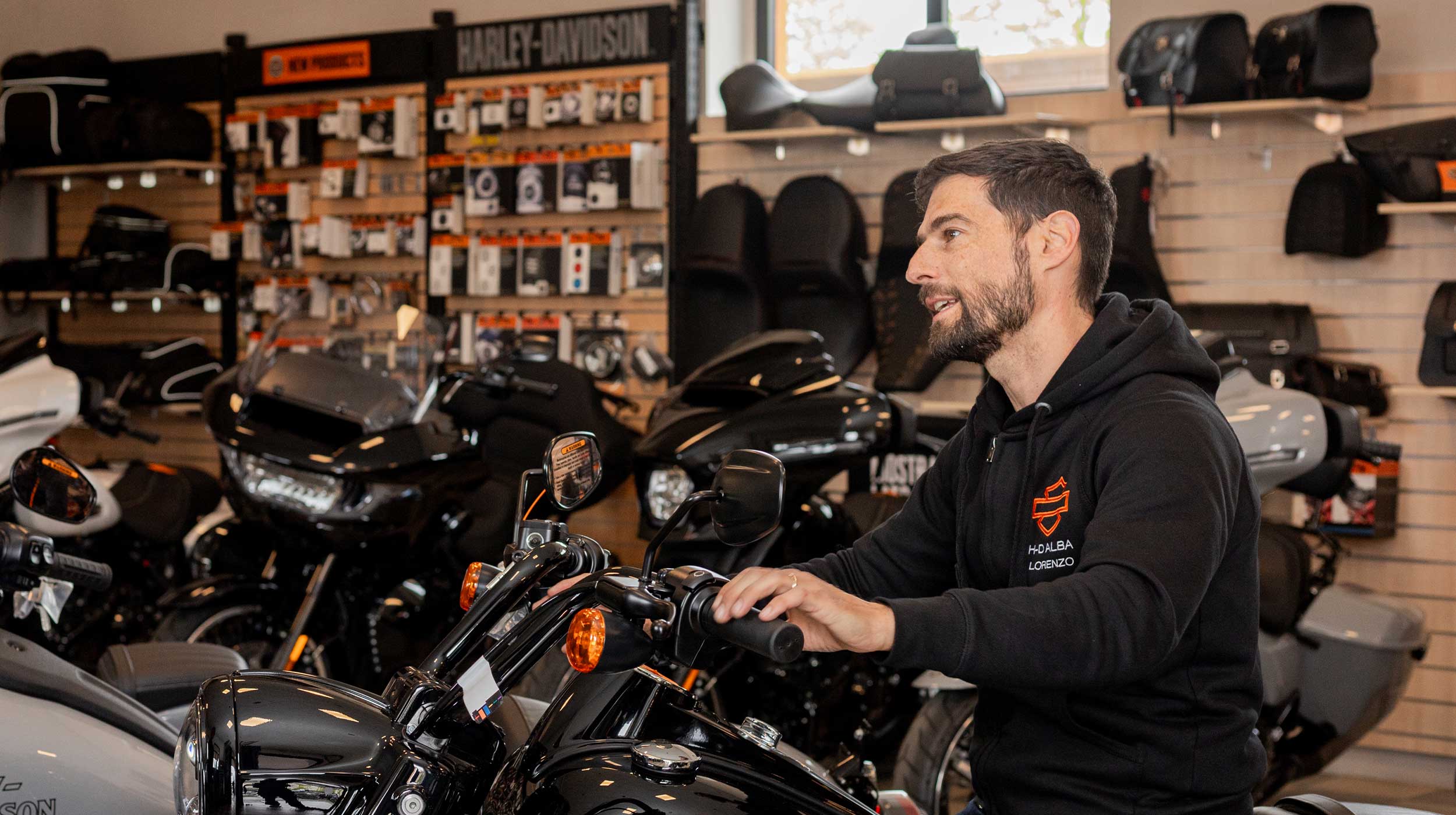 Esperienza Harley-Davidson: prova moto e abbigliamento H-DALBA Uomo con felpa Harley-Davidson H-DALBA Lorenzo su moto nera in showroom. Scaffali con accessori e abbigliamento moto sullo sfondo.
