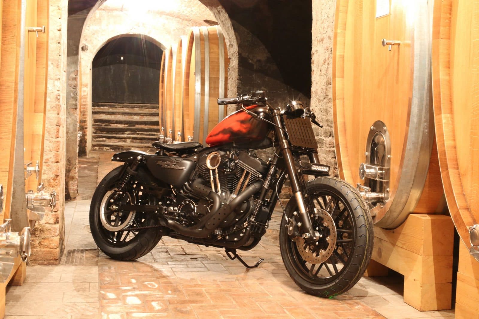 1484384897piccola2 Harley-Davidson customizzata, motore V-twin nero opaco, serbatoio rosso, forcella anteriore modificata, esposta in cantina tra botti di rovere e archi in mattoni.