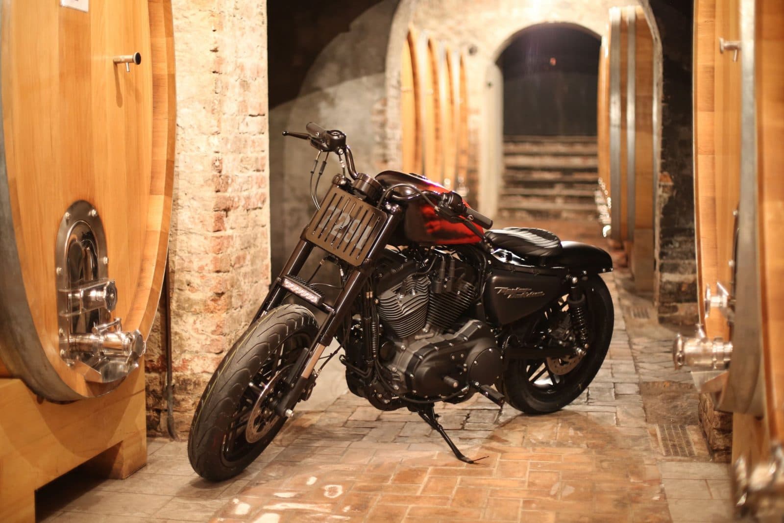 1484384912piccola4.jpg Motocicletta Harley-Davidson customizzata con serbatoio rosso e nero, parcheggiata in una cantina con botti di legno e pareti in mattoni a vista.