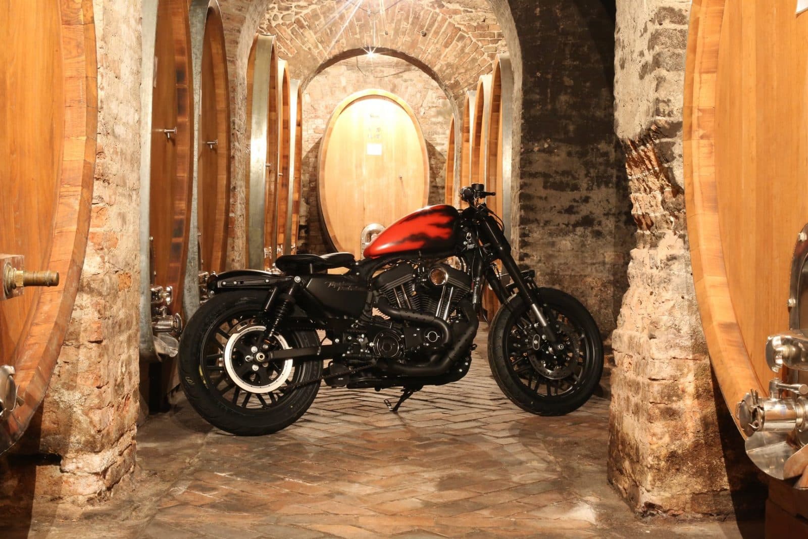 1484384937piccola1.jpg Moto Harley-Davidson Sportster customizzata con livrea fiammata, parcheggiata tra grandi botti di rovere in una cantina storica.