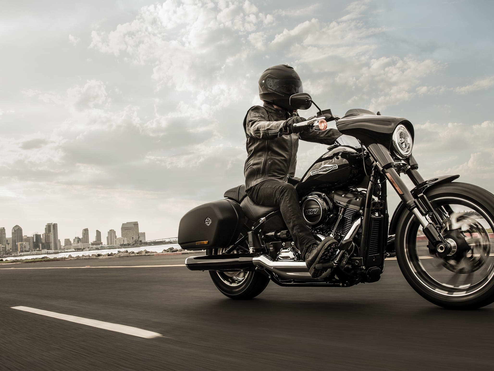 1511003456softail-scout-hdi-marquee Motociclista con abbigliamento tecnico e casco su Harley-Davidson nera, modello touring con borse laterali, in movimento su strada con skyline urbano.
