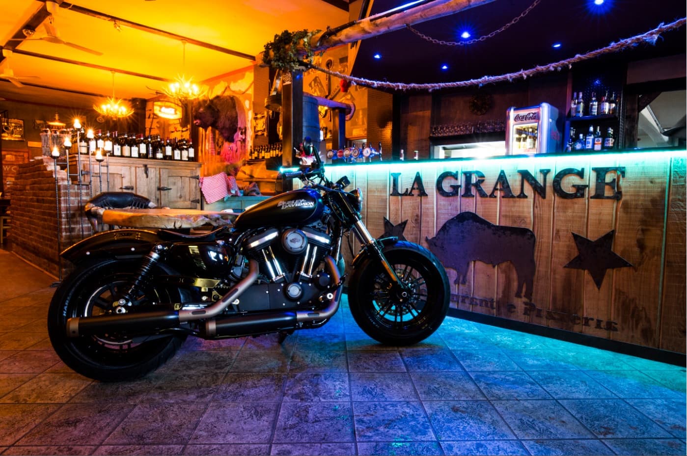 1516097276concorsobattle-1 Harley-Davidson nera in primo piano, parcheggiata in un locale rustico 'LA GRANGE'. L'illuminazione bicolore esalta la moto e l'ambiente.