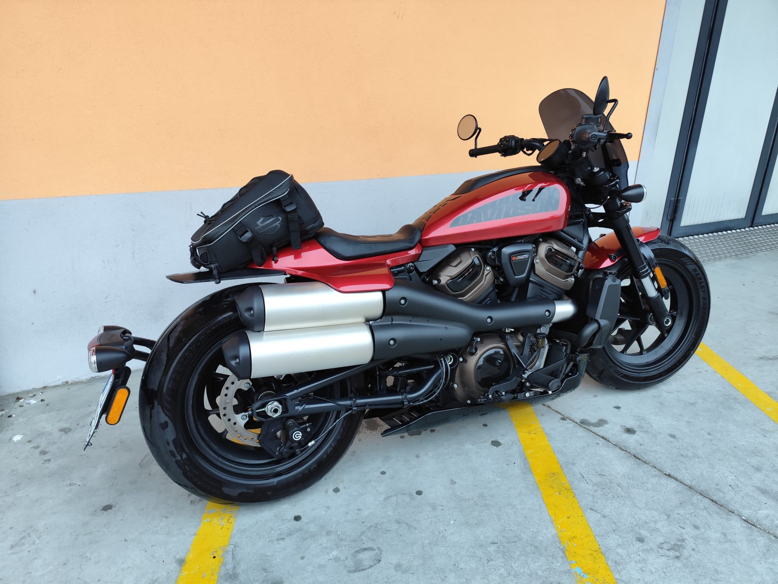 Harley-Davidson Sportster S: design e potenza per il tuo test ride. Harley-Davidson Sportster S rossa parcheggiata, con motore Revolution Max 1250T, scarico doppio, borsa posteriore e cupolino. Pneumatici bagnati su asfalto.