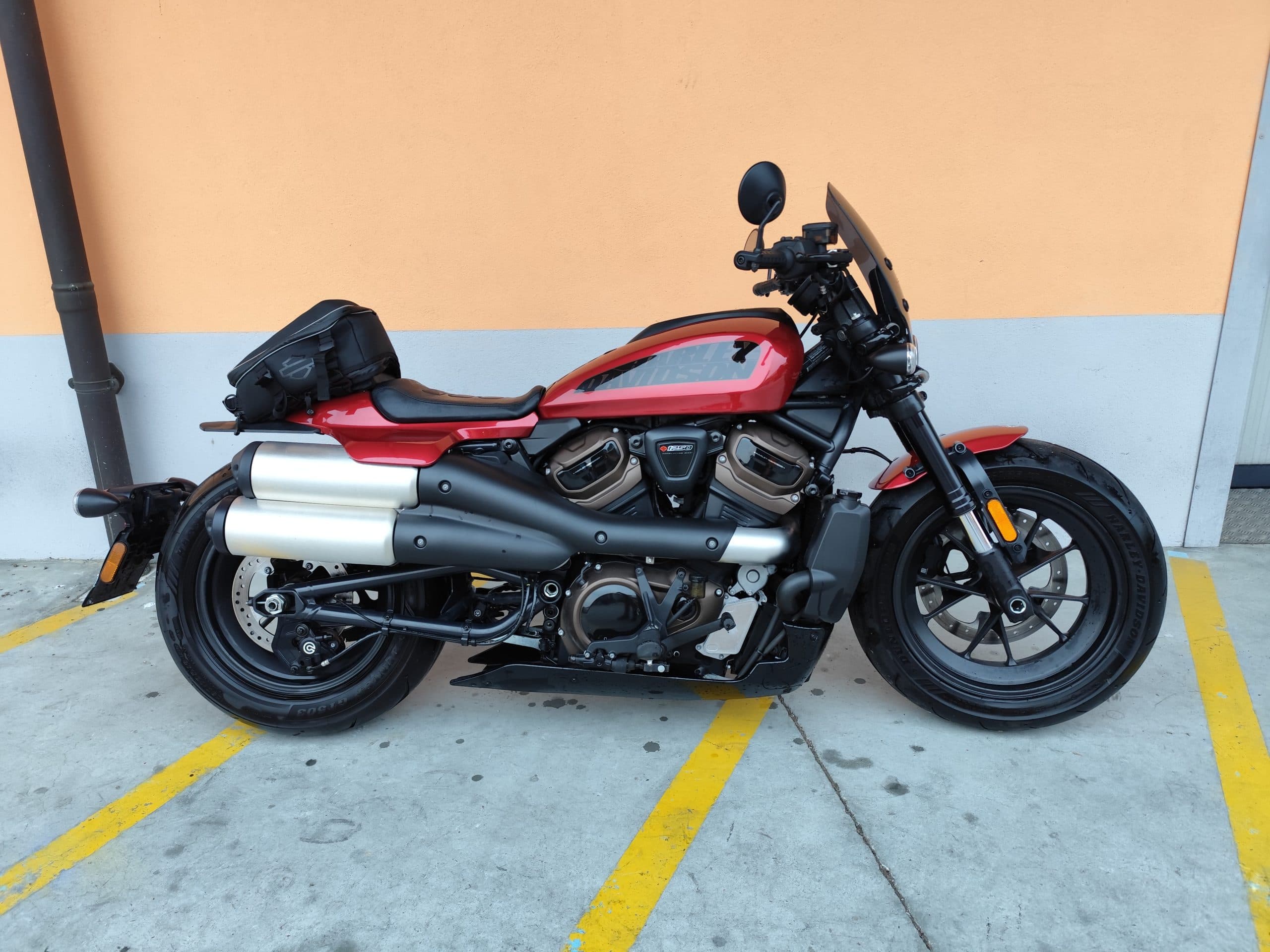 Harley-Davidson Sportster S rossa Motocicletta Harley-Davidson Sportster S rossa parcheggiata su asfalto con strisce gialle, affiancata da un muro bicolore arancione e grigio. La moto presenta un motore Revolution Max 1250T, scarichi alti cromati, una borsa da serbatoio nera e un cupolino fumé.