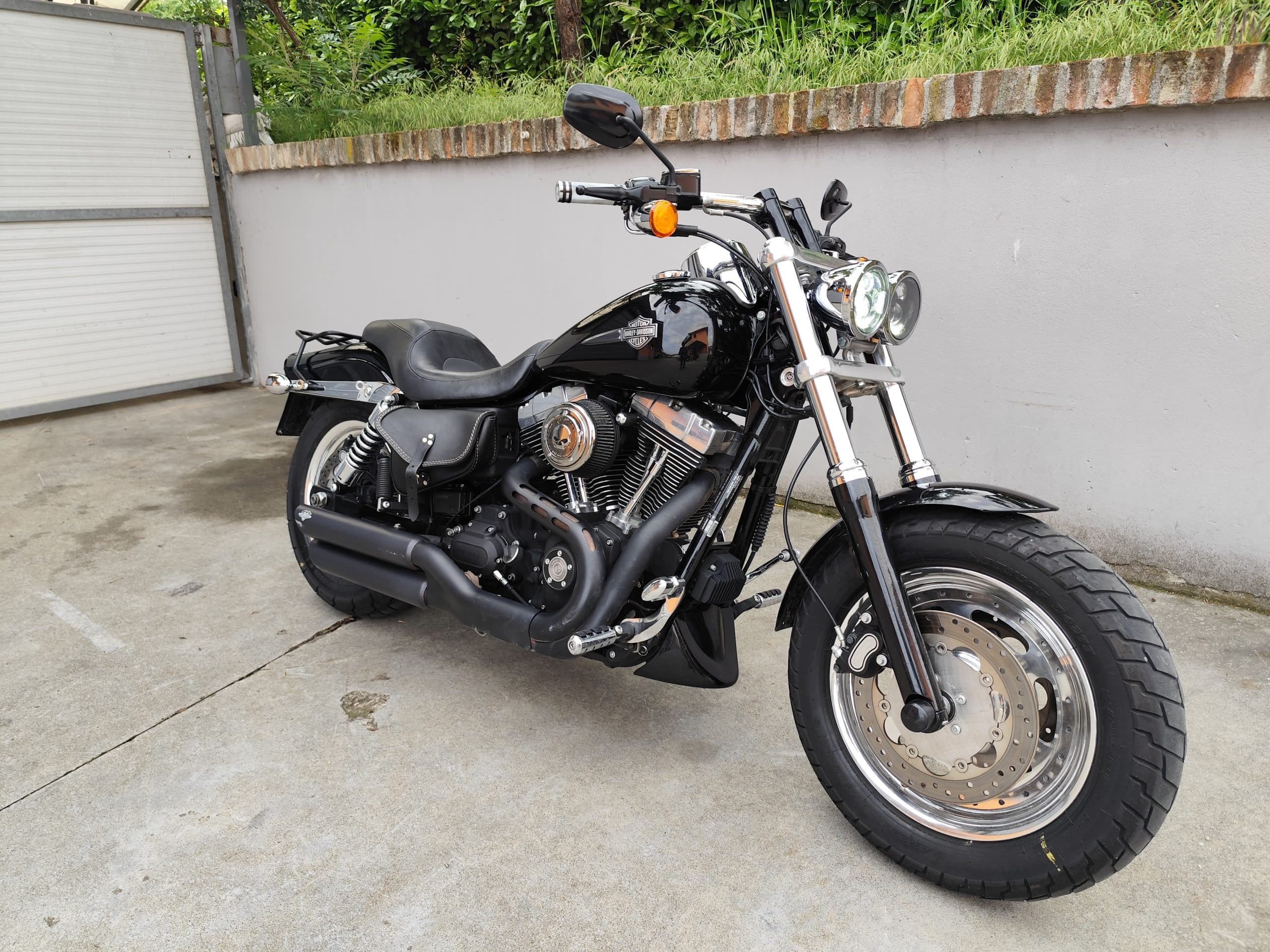 1749819270707.jpg Harley-Davidson Fat Bob nera lucida parcheggiata su cemento, con motore V-twin, scarichi neri opachi, borse laterali in pelle e cerchi cromati.