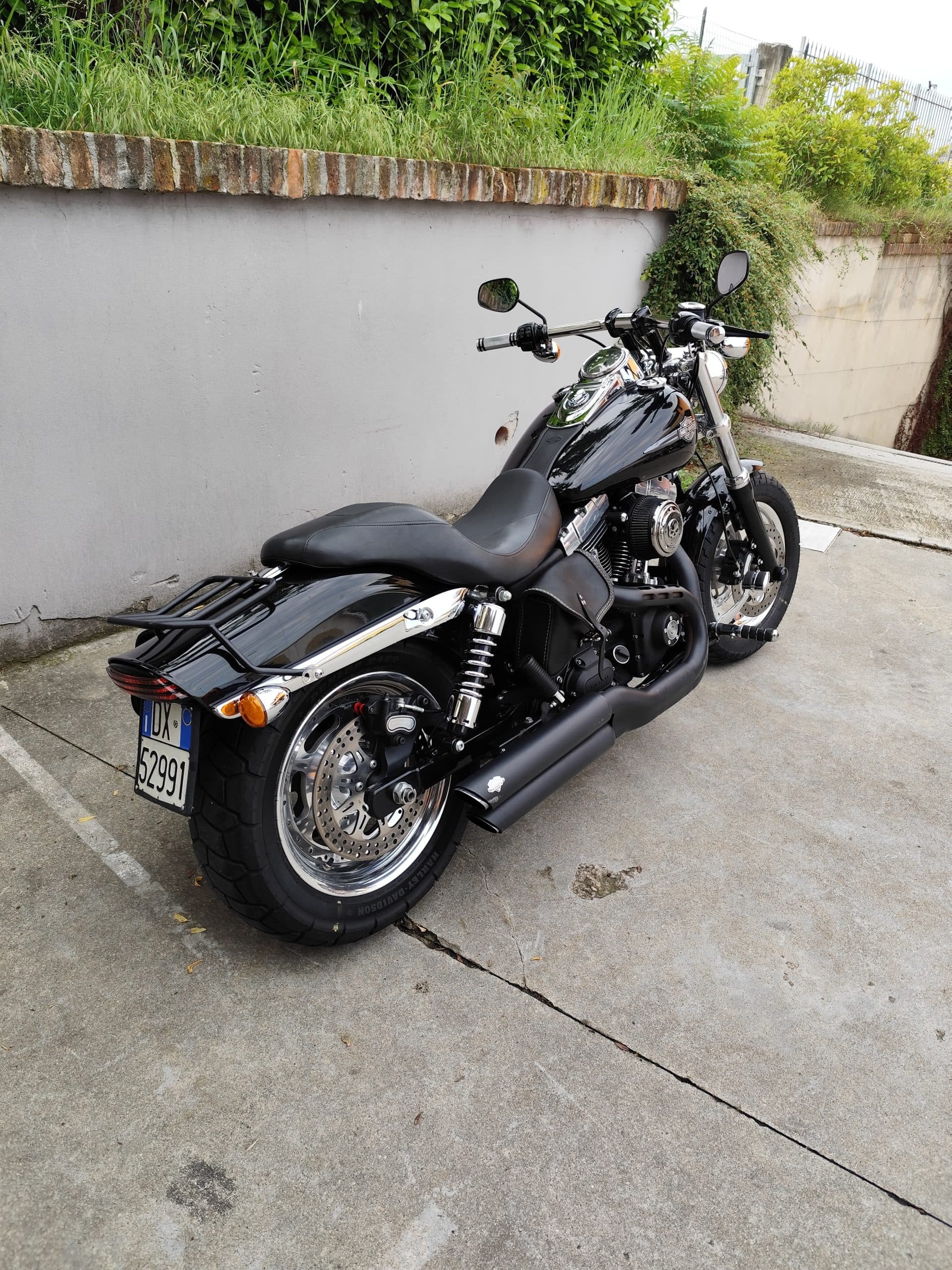 1749819270844.jpg Harley-Davidson nera, modello cruiser, parcheggiata su pavimentazione in cemento. Dettagli di scarichi neri, cerchi cromati e portapacchi posteriore.