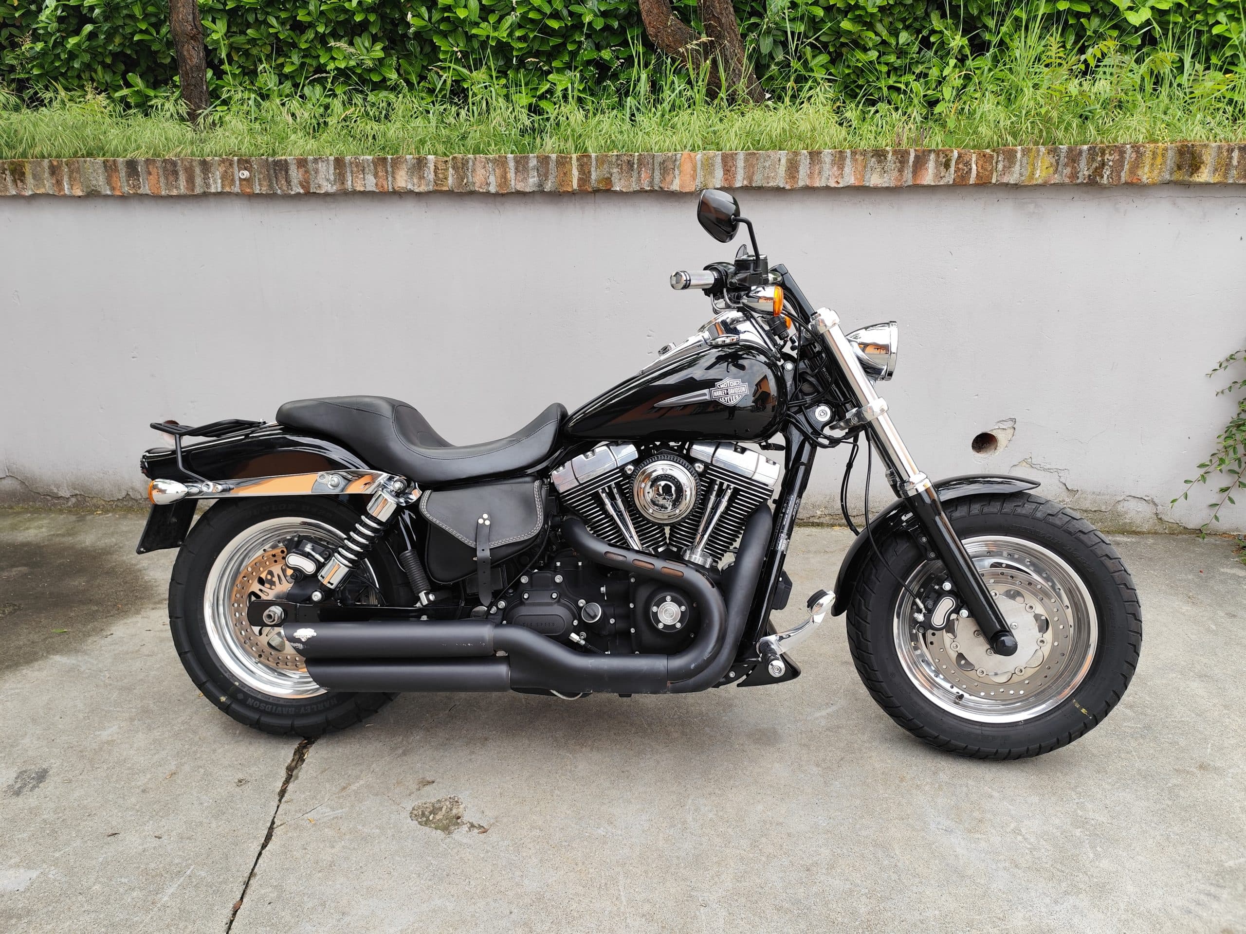 1749819270935-scaled Moto Harley-Davidson nera, stile cruiser, con pneumatici larghi, motore cromato e scarichi doppi, esposta su pavimentazione in cemento.