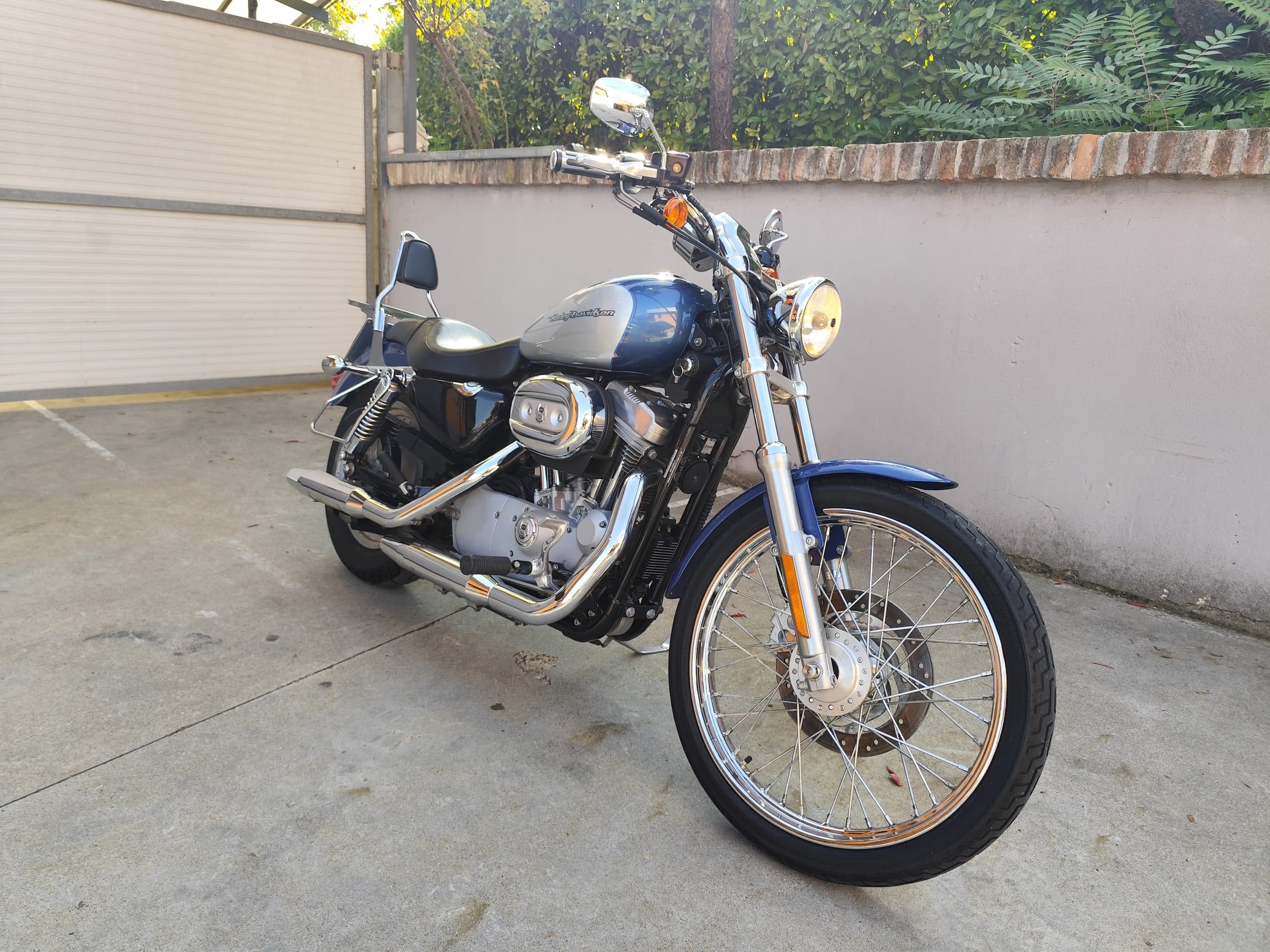 1758557801306.jpg Harley-Davidson Sportster blu e argento, con motore V-twin e scarichi cromati, parcheggiata. Dettagli ruote a raggi e faro acceso.