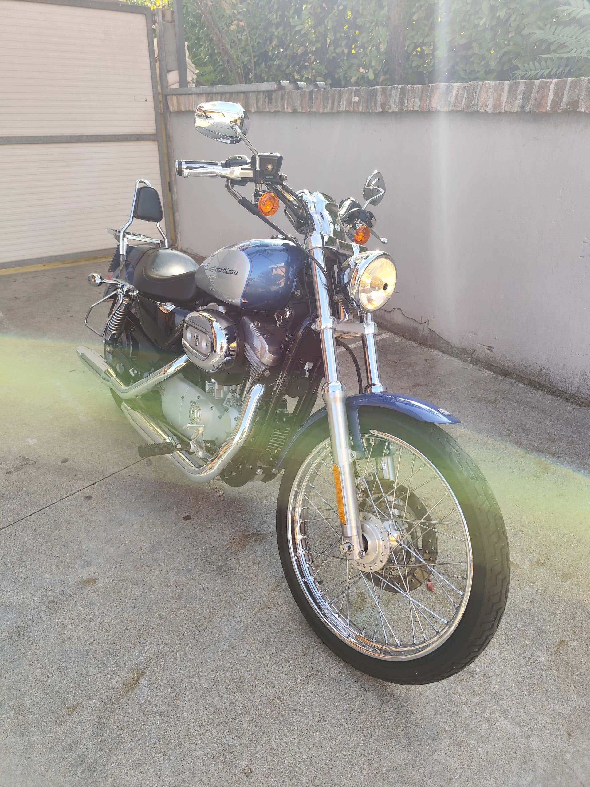 1758557801339.jpg Moto Harley-Davidson Sportster bicolore blu e argento, motore V-Twin, cerchi a raggi cromati e faro acceso, su pavimentazione esterna.