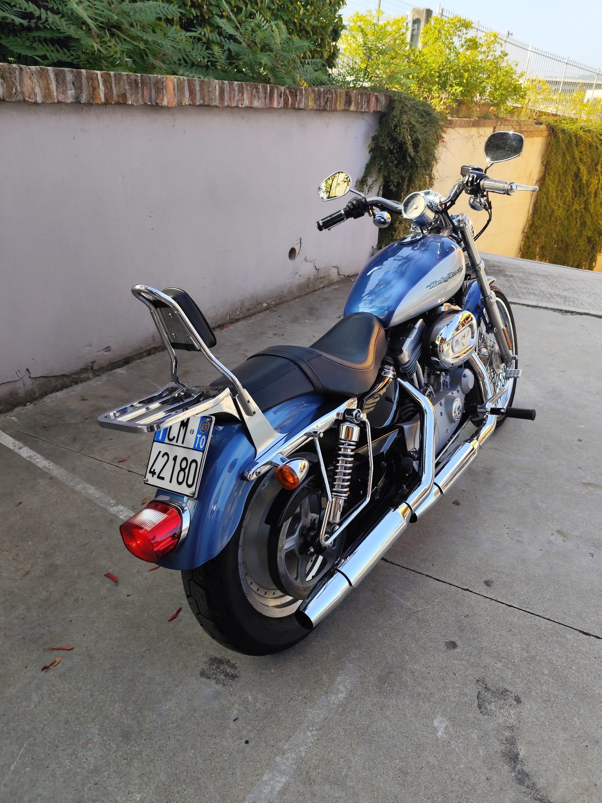 1758557801415.jpg Harley-Davidson Sportster blu e argento parcheggiata, con dettagli cromati lucidi, scarichi, motore e portapacchi. Ideale per test ride.