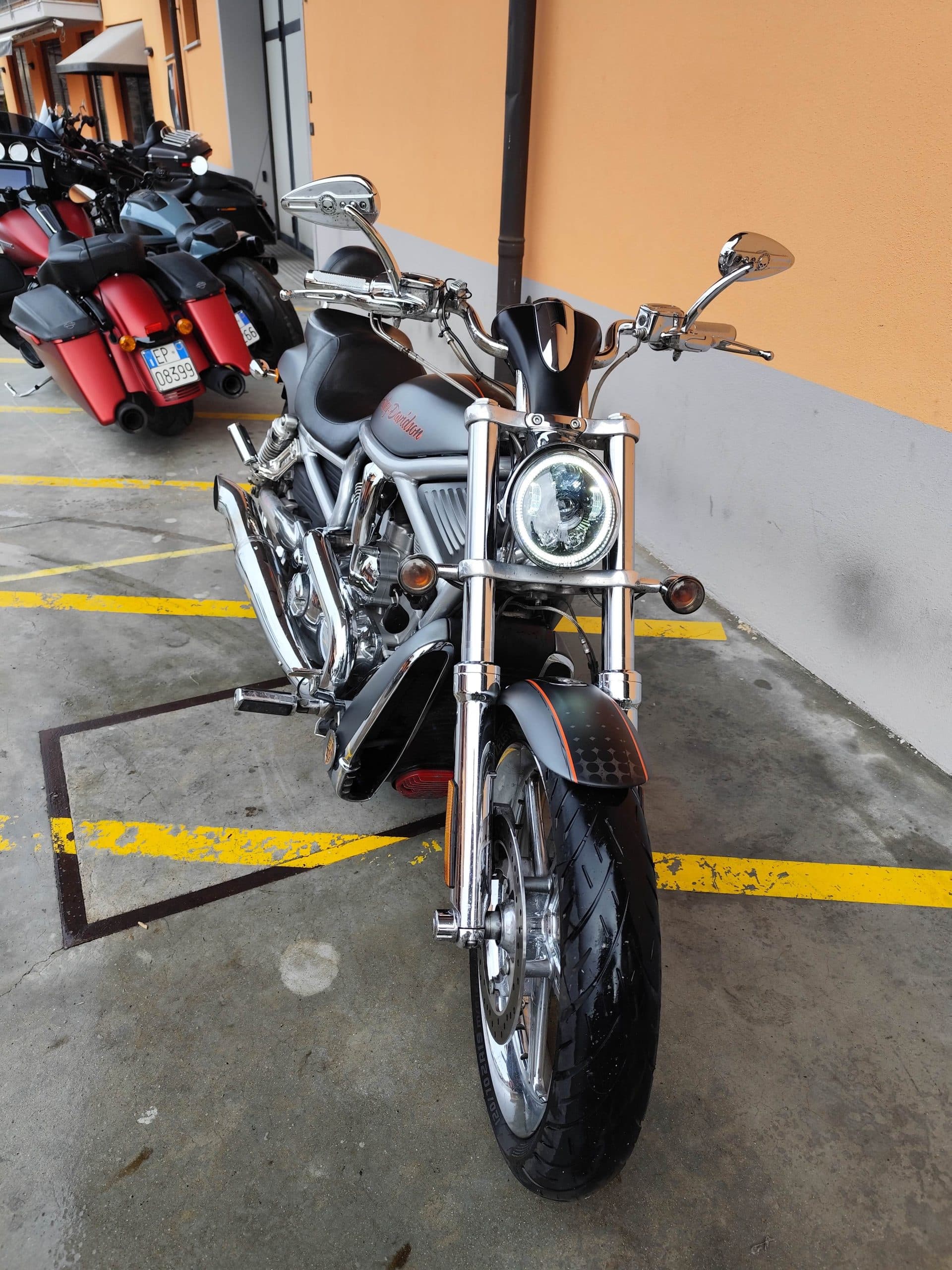 1766134081816.jpg Harley-Davidson V-Rod grigia con faro LED, forcelle e scarichi cromati, in primo piano. Sullo sfondo, moto touring Harley-Davidson rosse.