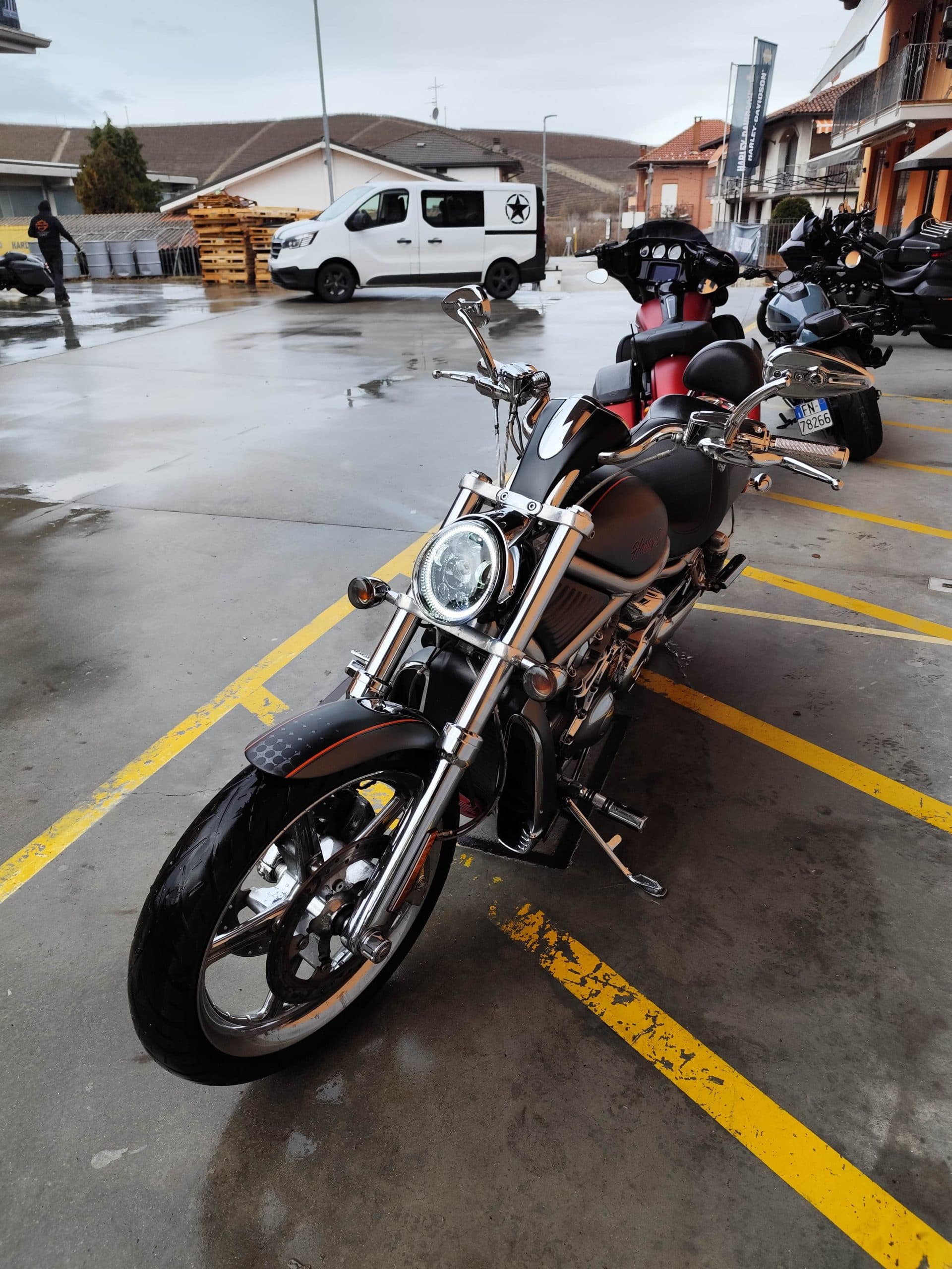 1766134081867.jpg Moto Harley-Davidson customizzata nera con finiture cromate e faro LED circolare, parcheggiata su asfalto bagnato. Sfondo con altre moto e banner Harley-Davidson.