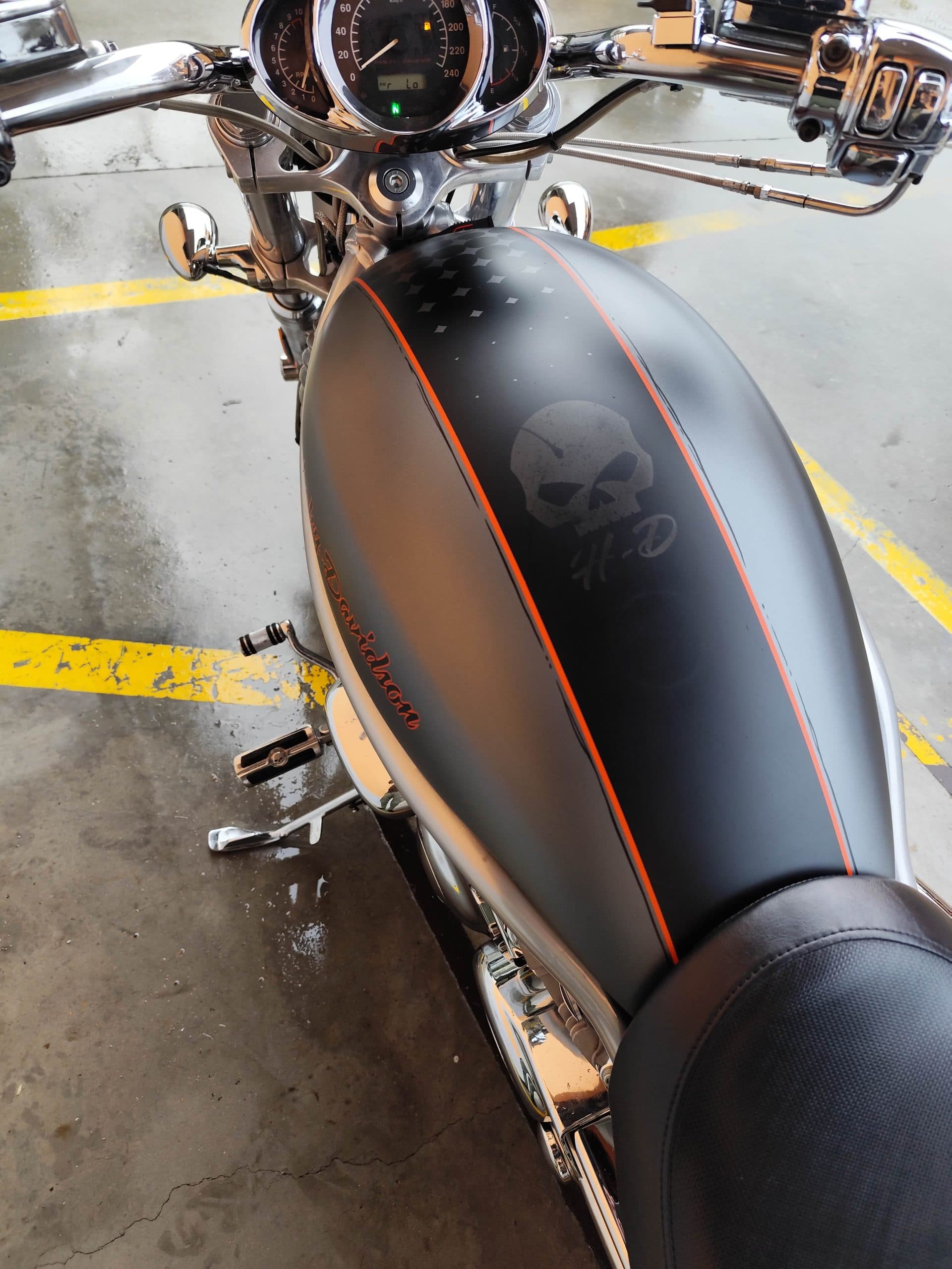 1766134081920.jpg Vista dall'alto di una moto Harley-Davidson, serbatoio nero opaco con strisce arancioni e teschio H-D. Manubrio crom