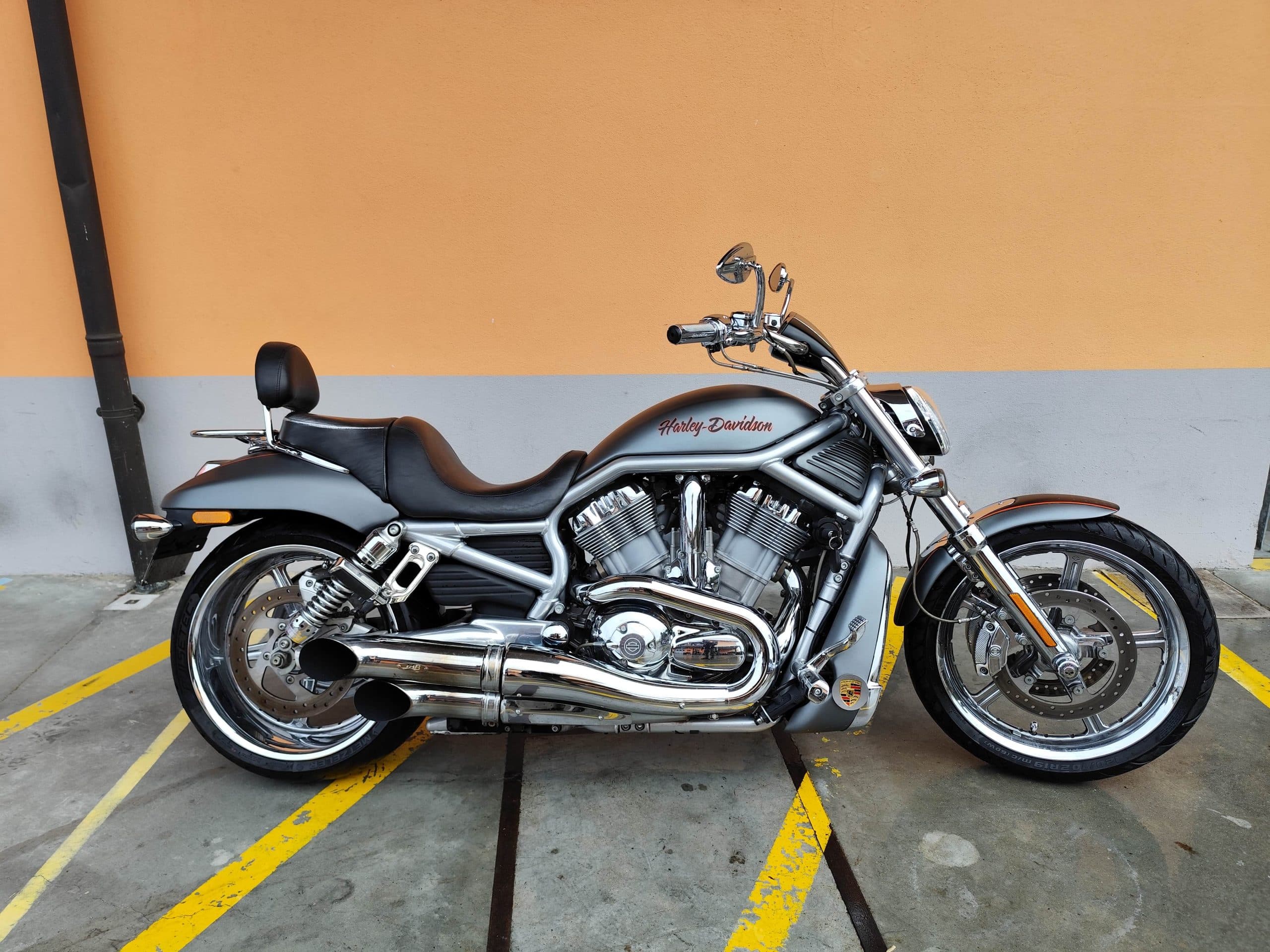 1766134082251-scaled Harley-Davidson V-Rod grigia con dettagli cromati, parcheggiata su asfalto con segnaletica gialla, sfondo parete arancione. Ideale per test ride.