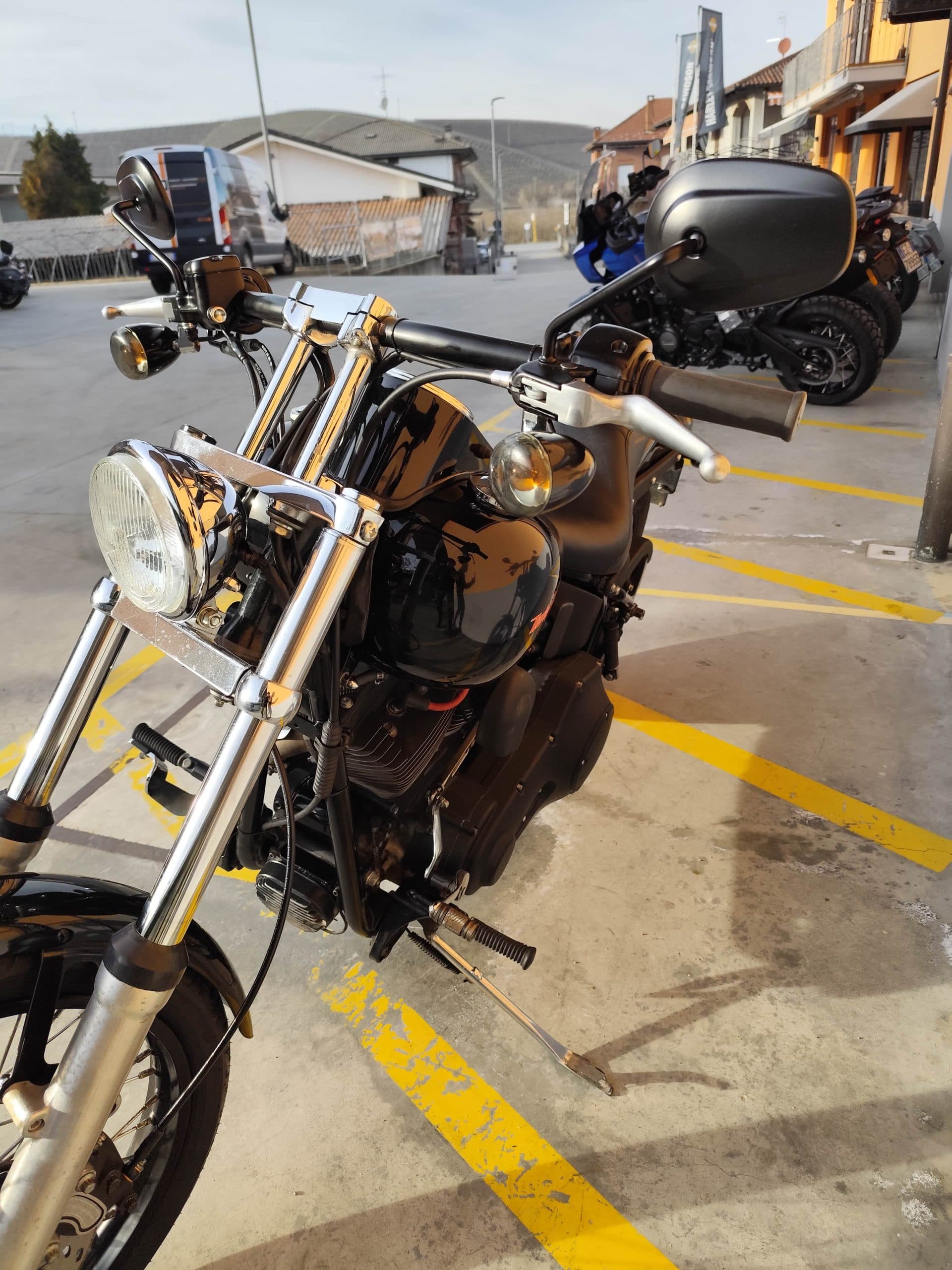 1768227369402.jpg Harley-Davidson nera parcheggiata in concessionaria, con dettagli cromati su manubrio, faro e forcelle. Altre moto e bandiere Harley-Davidson sullo sfondo.