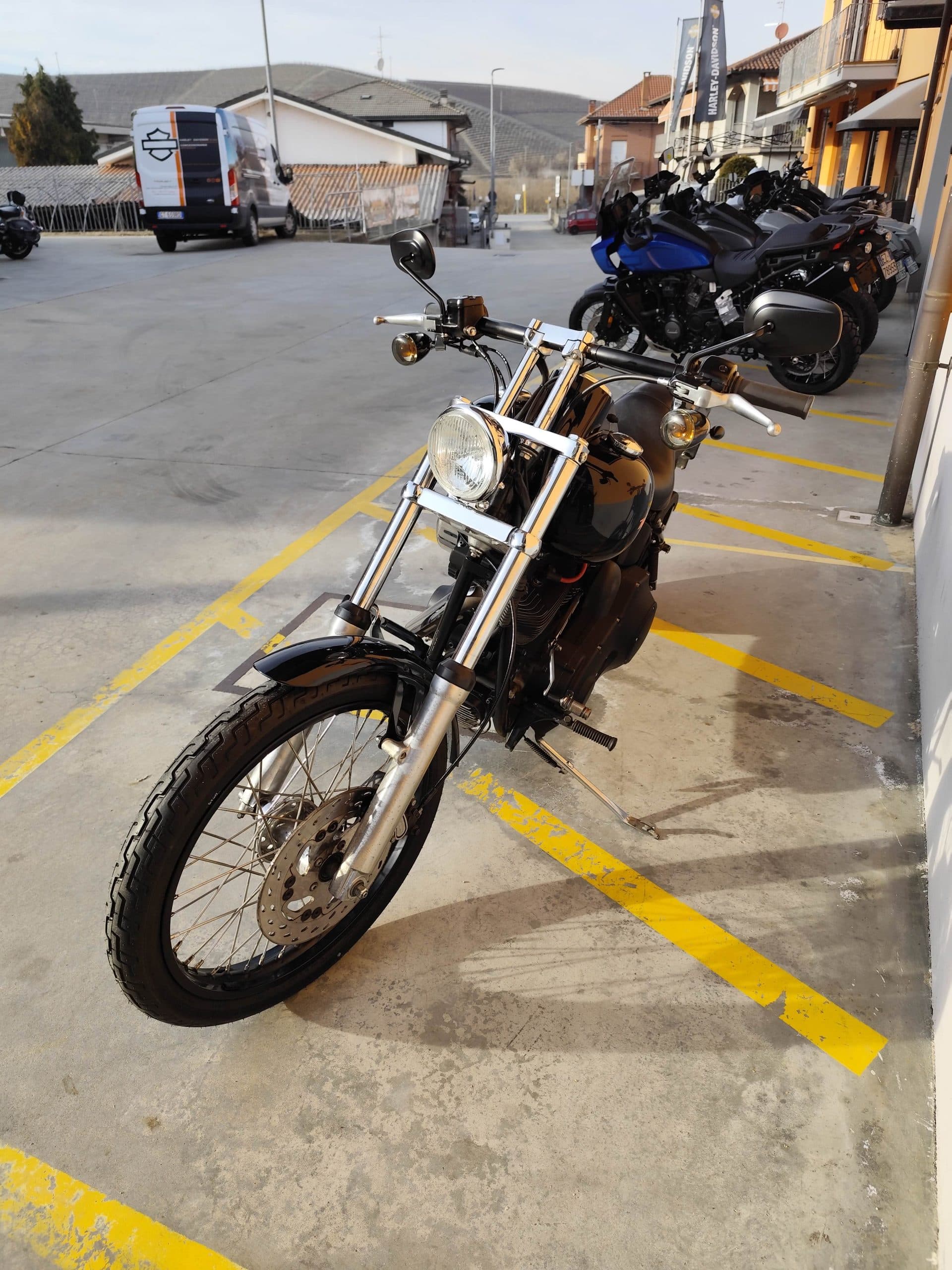 1768227369463.jpg Harley-Davidson nera, stile chopper, con forcelle cromate e faro tondo, parcheggiata nel piazzale di una concessionaria. Altre moto e furgone Harley-Davidson sullo sfondo, con vigneti.