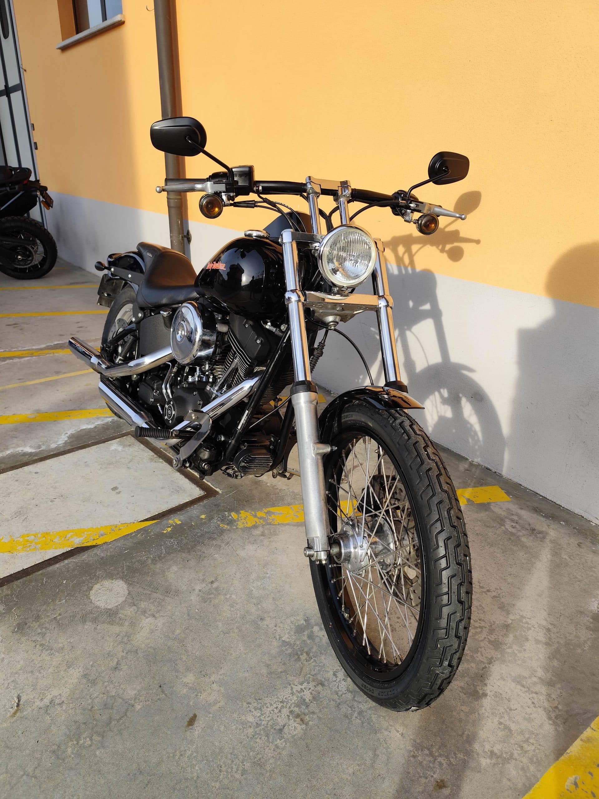 1768227369822.jpg Moto Harley-Davidson nera lucida con forcelle e scarichi cromati, ruota a raggi, parcheggiata all'aperto sotto il sole.