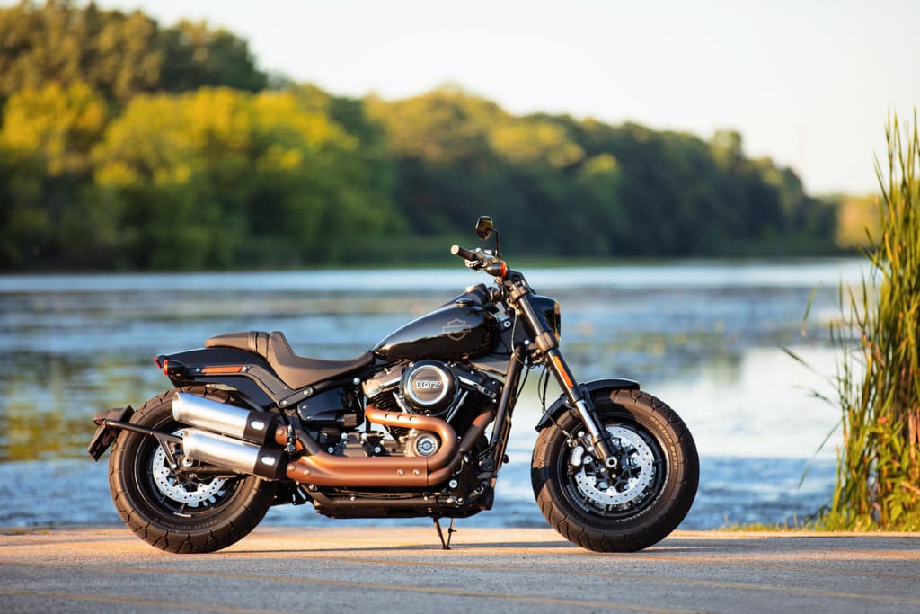 201461_my21_fxfbs_beauty_hdi_0640_jk.jpg Harley-Davidson Fat Bob nera, motore 107, scarichi bronzo, parcheggiata su asfalto vicino a un lago.