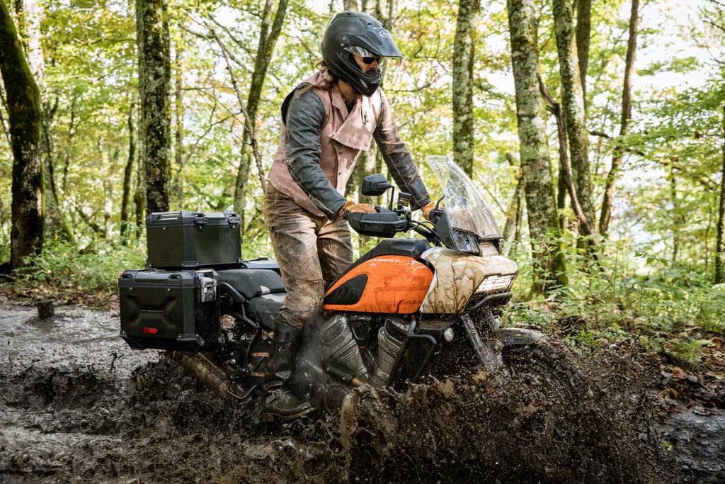 21-Pan-America-Momoa.jpg Motociclista con abbigliamento tecnico su Harley-Davidson Pan America in un test ride off-road, affronta un percorso fangoso nel bosco.