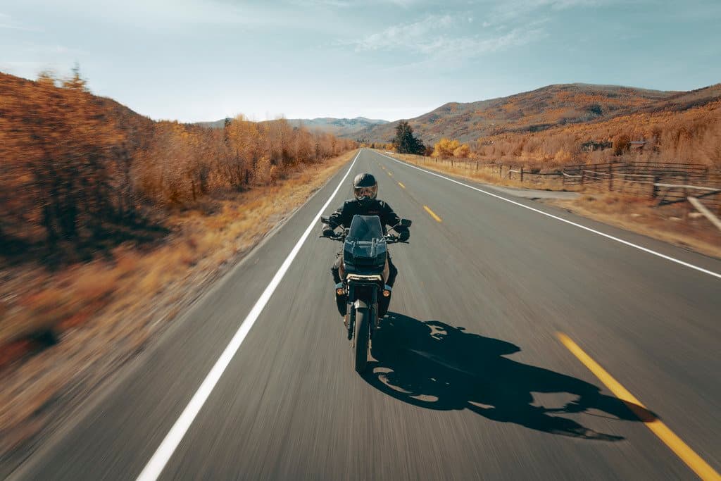 Harley-Davidson Pan America: test ride e avventura su strada. Motociclista con abbigliamento tecnico e casco guida una Harley-Davidson Pan America su strada asfaltata tra colline con foliage autunnale.