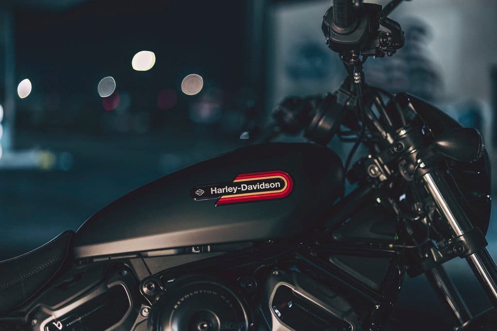 23-rh975s-nightsterspecial-detail-ab-0001.jpg Dettaglio notturno del serbatoio di una moto Harley-Davidson, con logo e strisce rosse e gialle, e luci bokeh sullo sfondo.