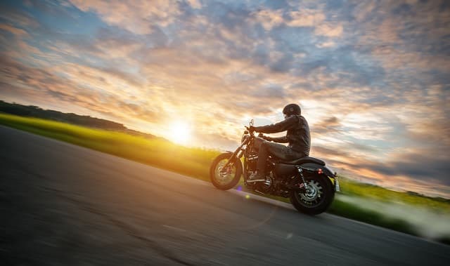 AdobeStock_157417016_ok Motociclista con abbigliamento tecnico in sella a una Harley-Davidson in movimento su strada, con sole al tramonto e sfondo sfocato.