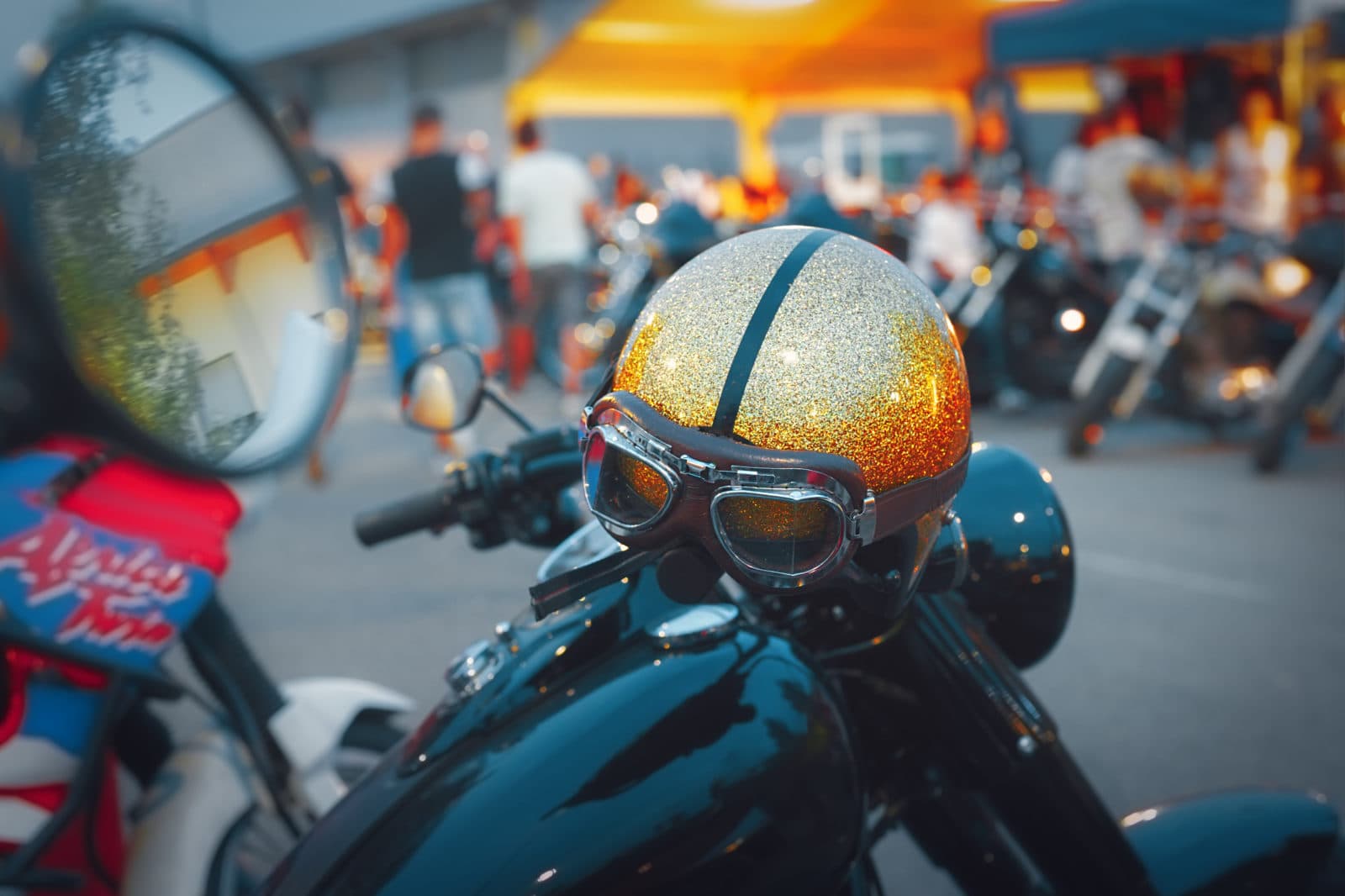 AdobeStock_220683398 Dettaglio di casco da moto glitterato oro e argento con occhiali vintage, poggiato su una moto in un evento Harley-Davidson.
